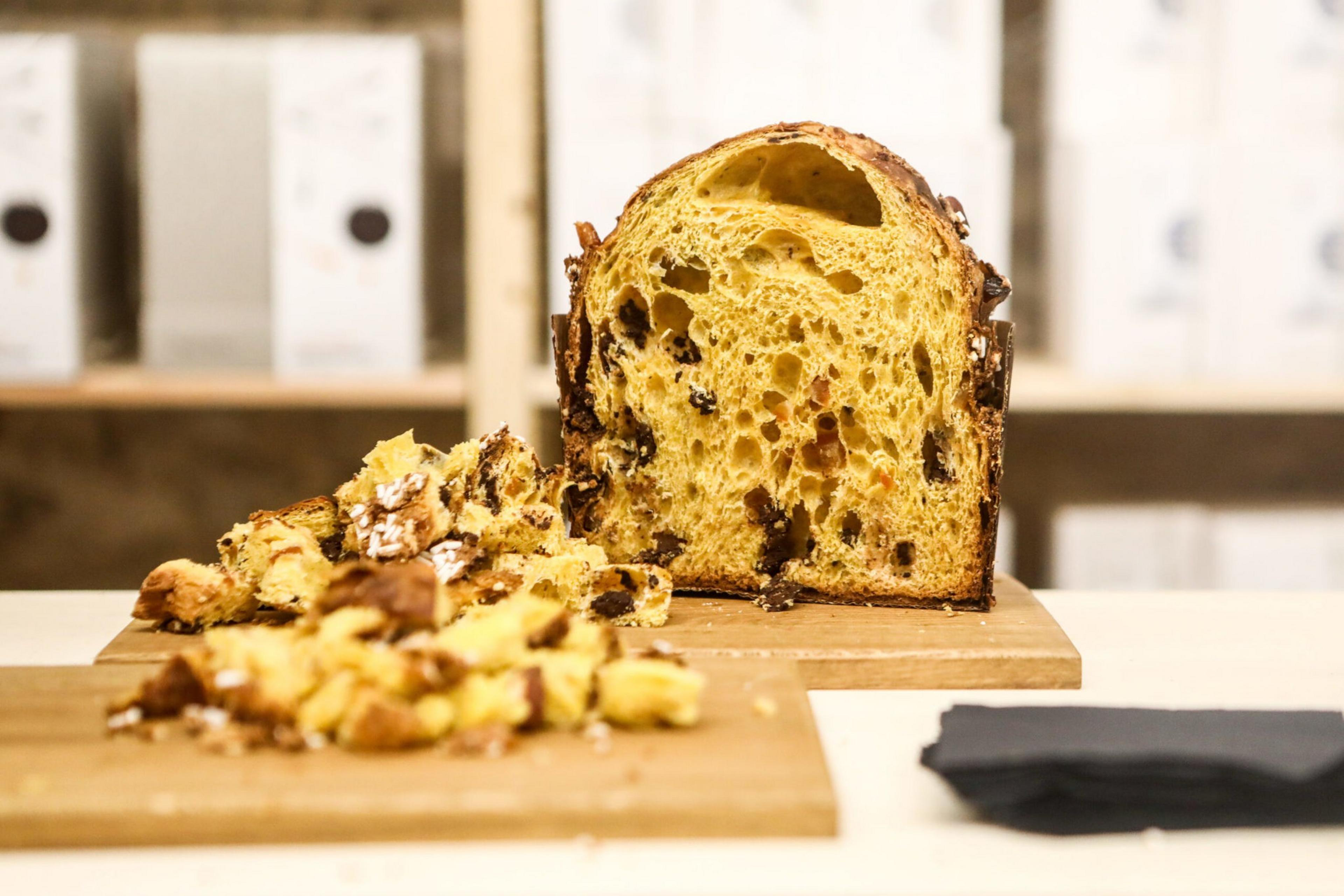 A sliced panettone with dried fruit sits on a wooden board, with crumbs scattered in front and a folded dark napkin to the right.