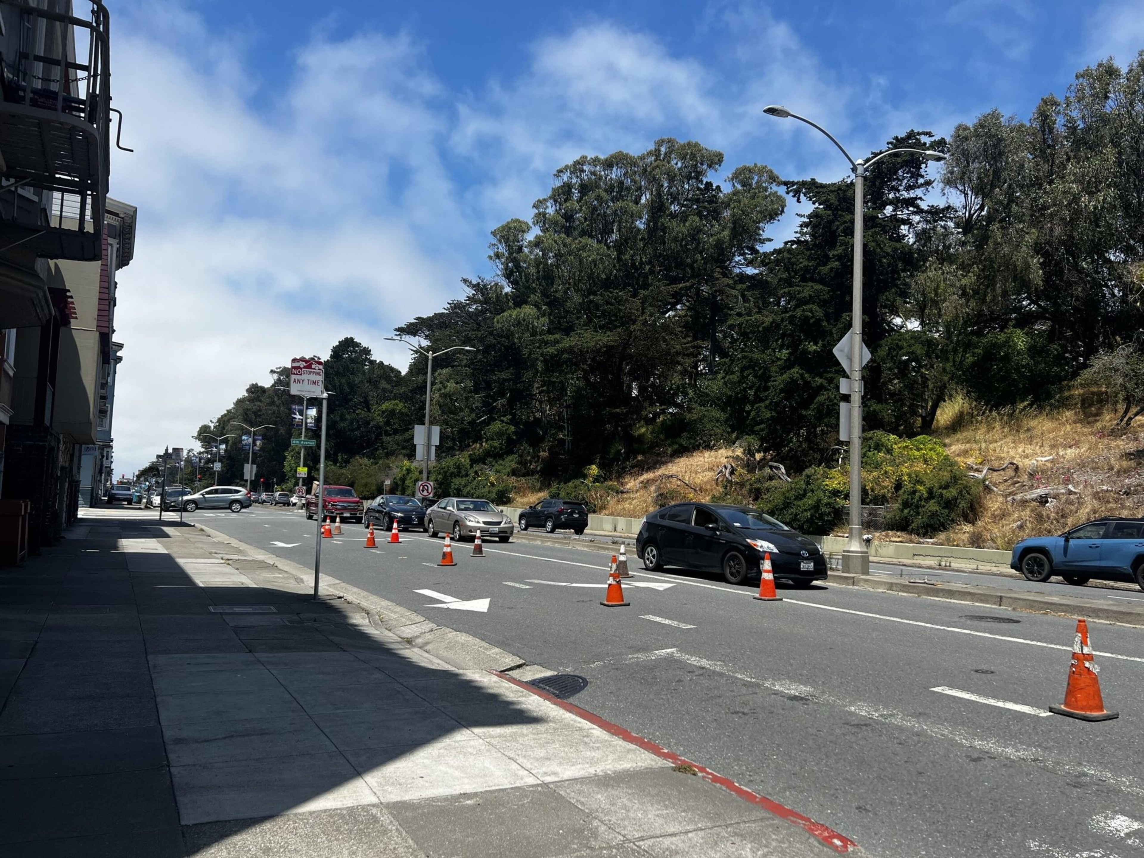 A street with traffic cones separating lanes, several cars waiting in line, a sidewalk on the left, and trees with dry grass on the right under a partly cloudy sky.