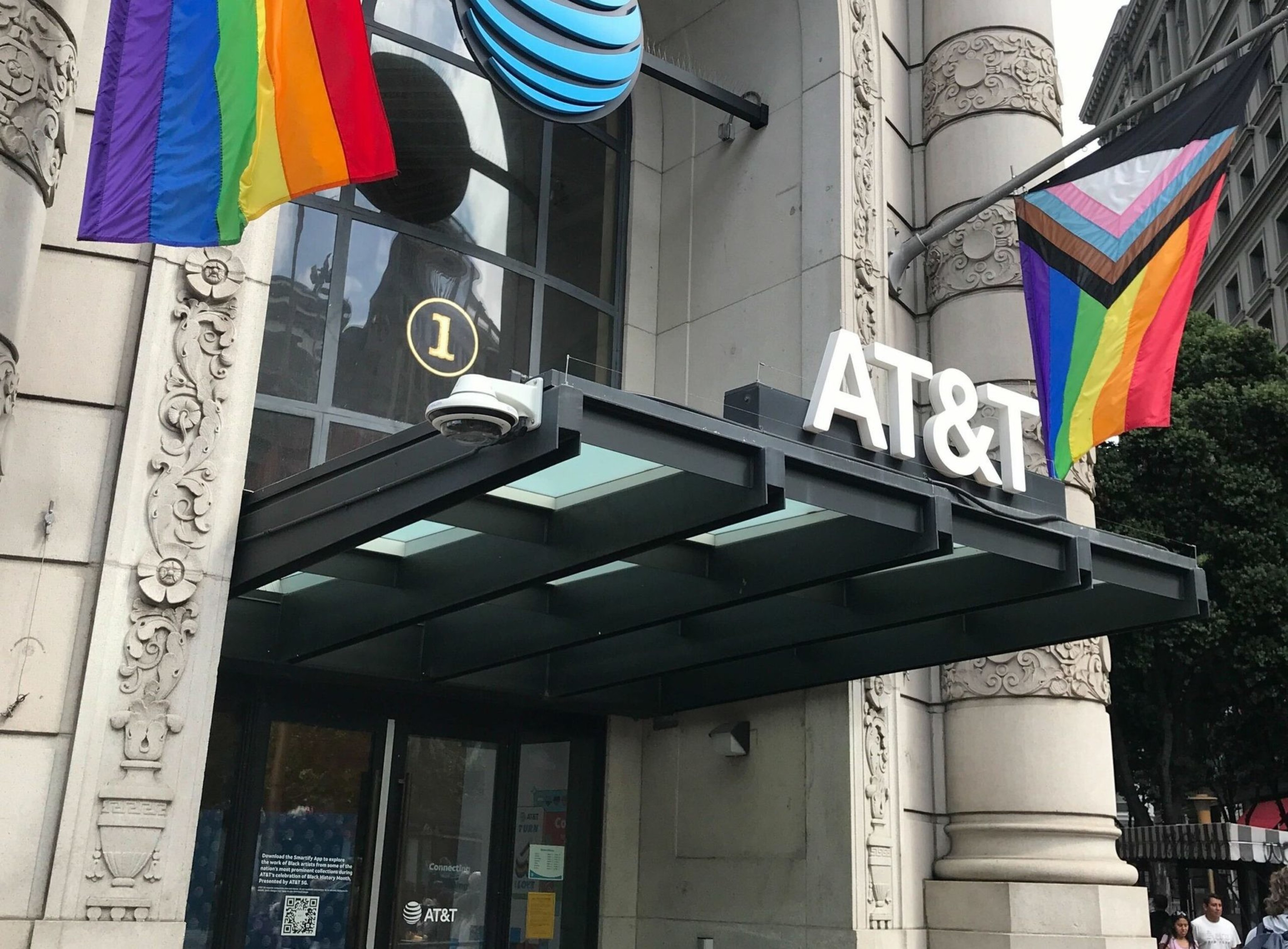AT&T store entrance with a glass canopy, LGBTQ+ rainbow and progress pride flags, ornate stone columns, and a security camera above the door.