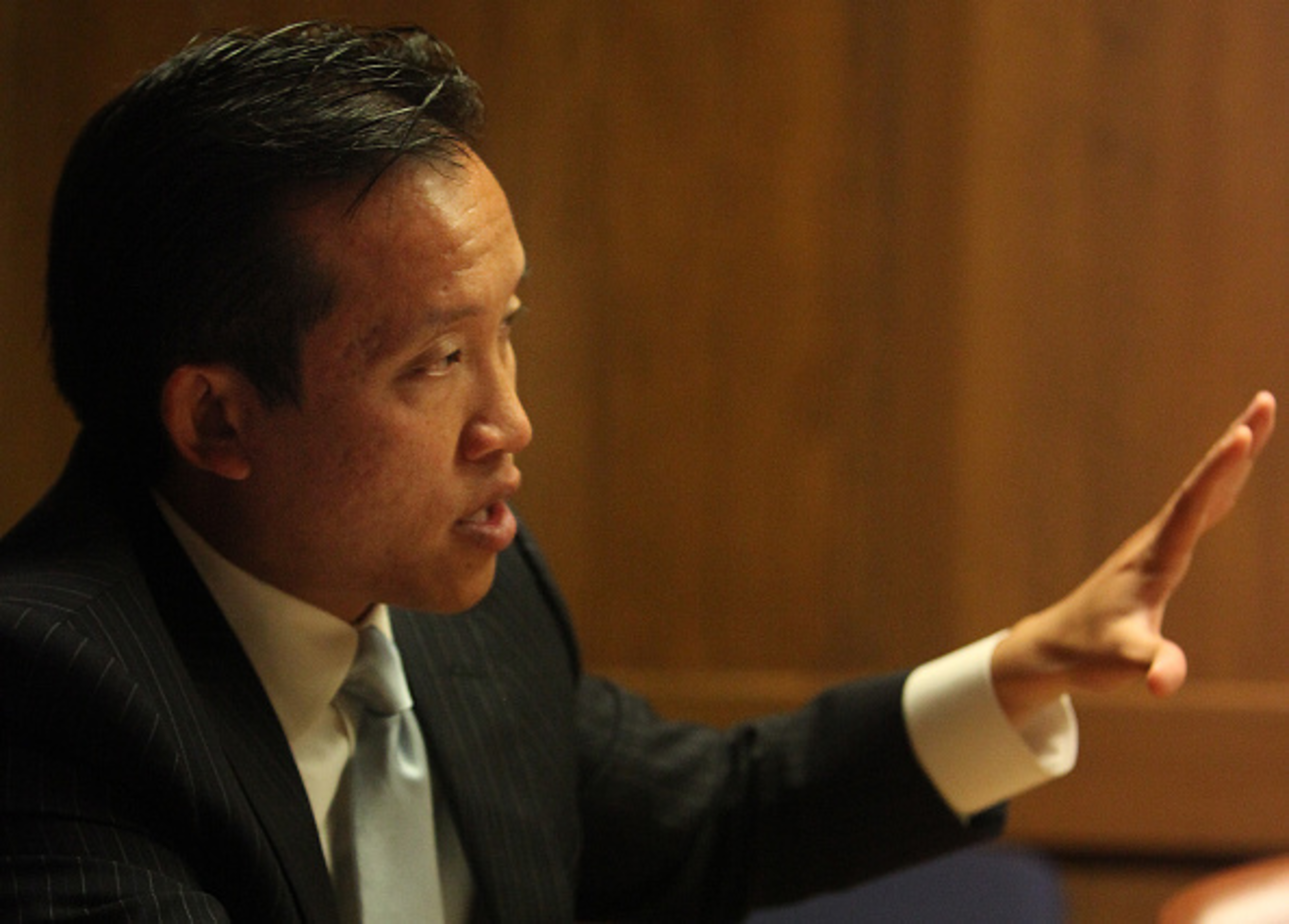 A man in a pinstripe suit and light blue tie gestures with his right hand while speaking, against a wooden background.