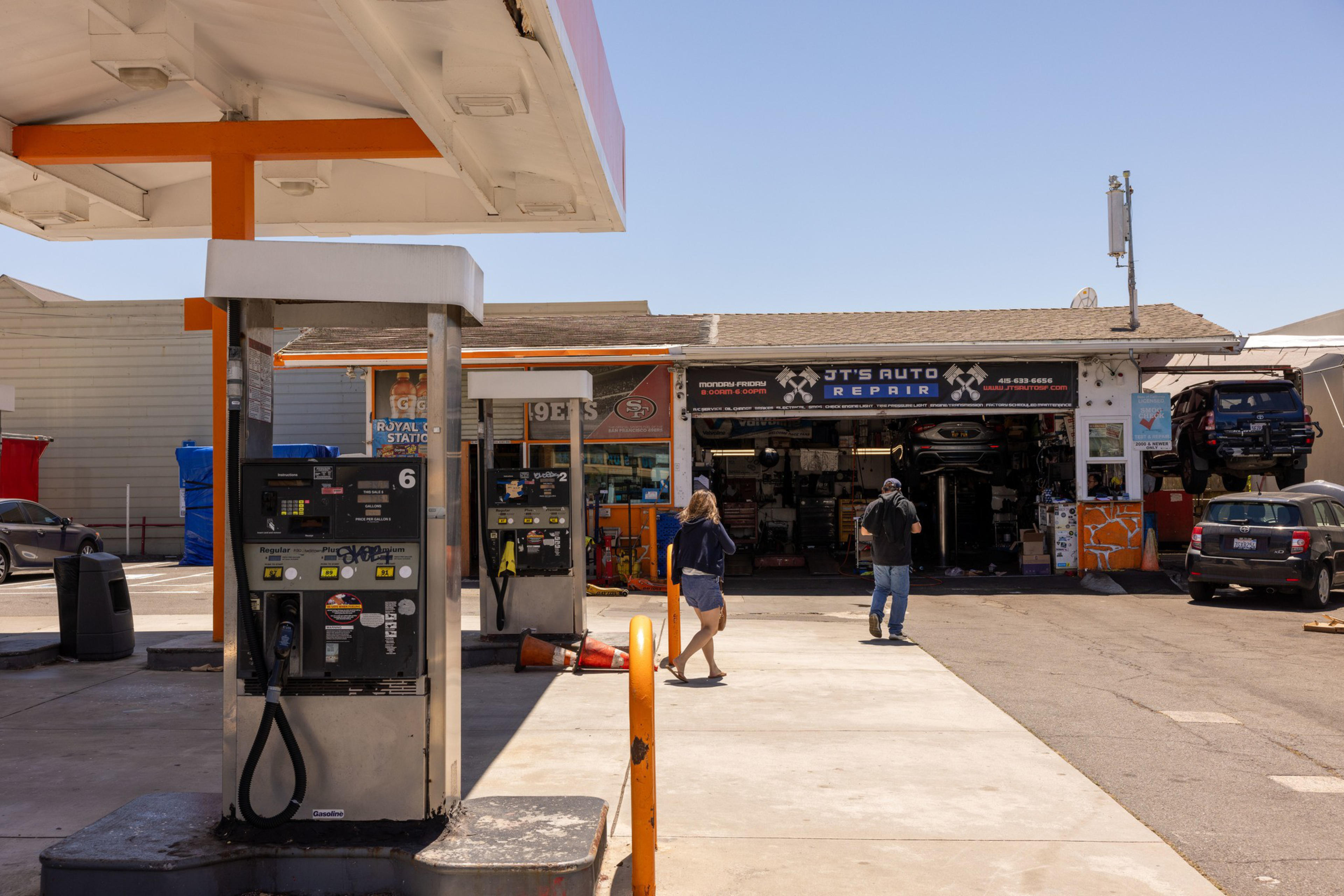 A gas station with a convenience store and an auto repair shop in the background. Two people walk toward the auto repair area, where a car is on a lift.