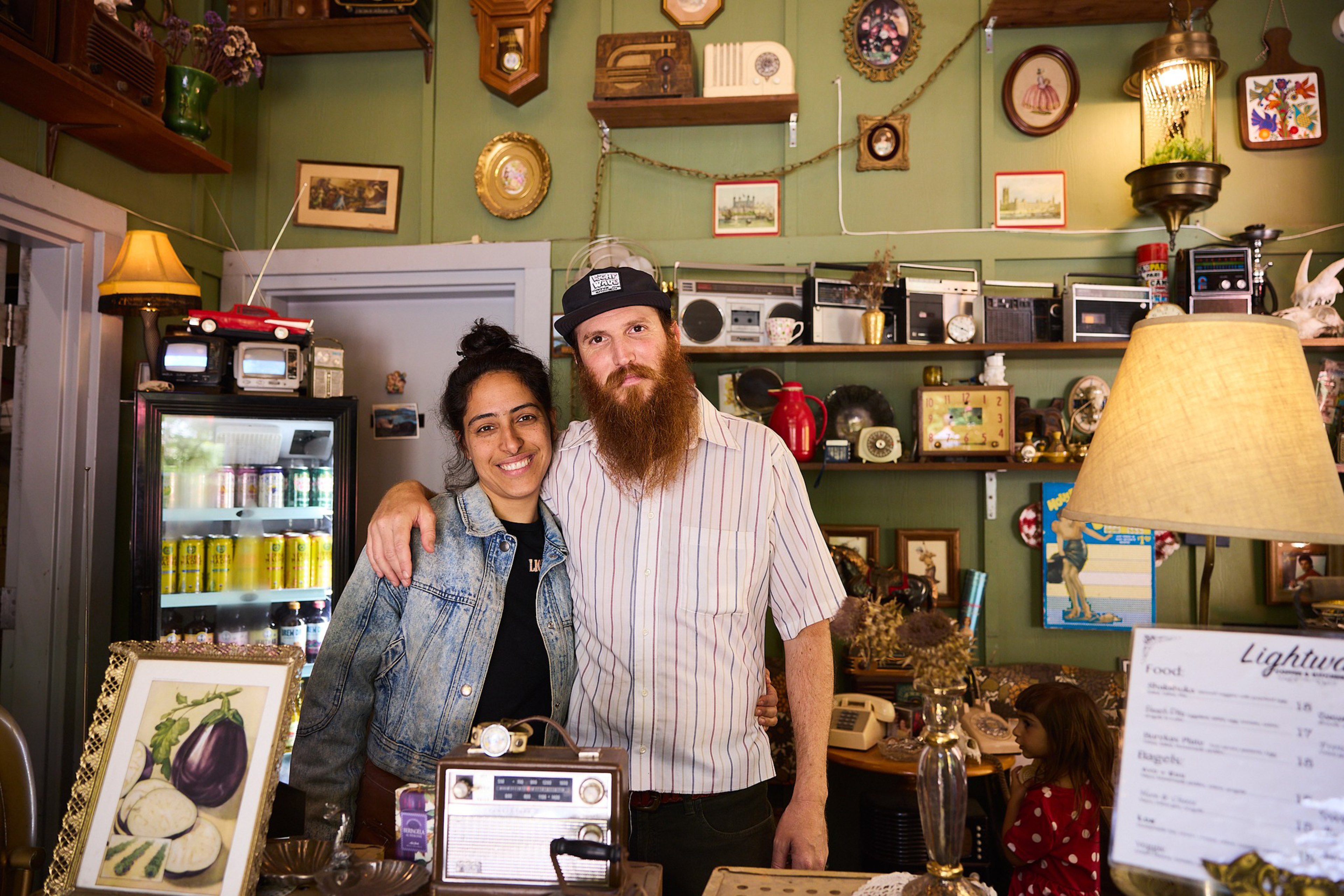 A man with a long beard and a woman smile, standing close in a cozy, vintage-style room decorated with old radios, framed pictures, and a lamp.
