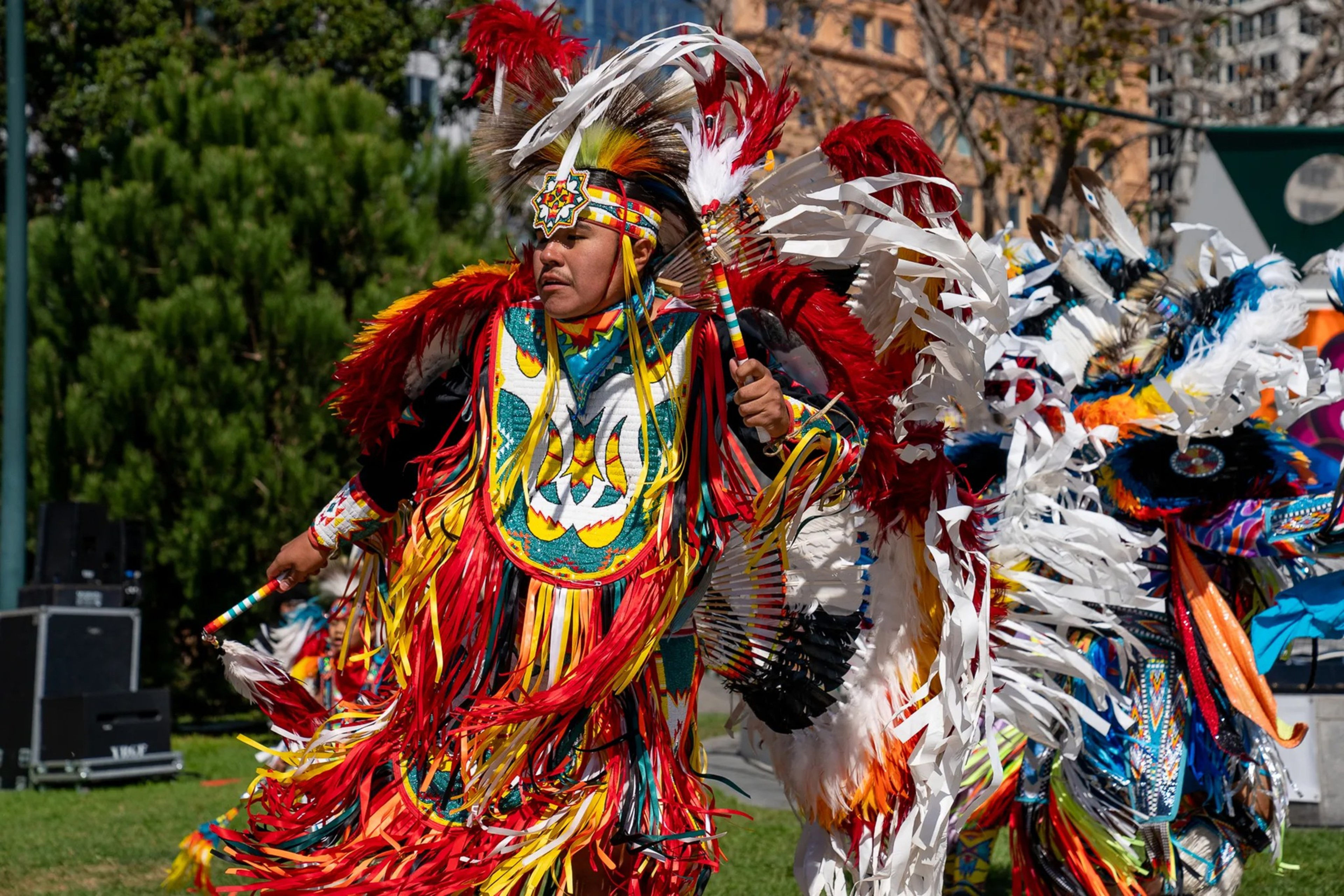 A person wears vibrant red, yellow, and white feathered regalia decorated with patterns, dancing outdoors during a cultural celebration.