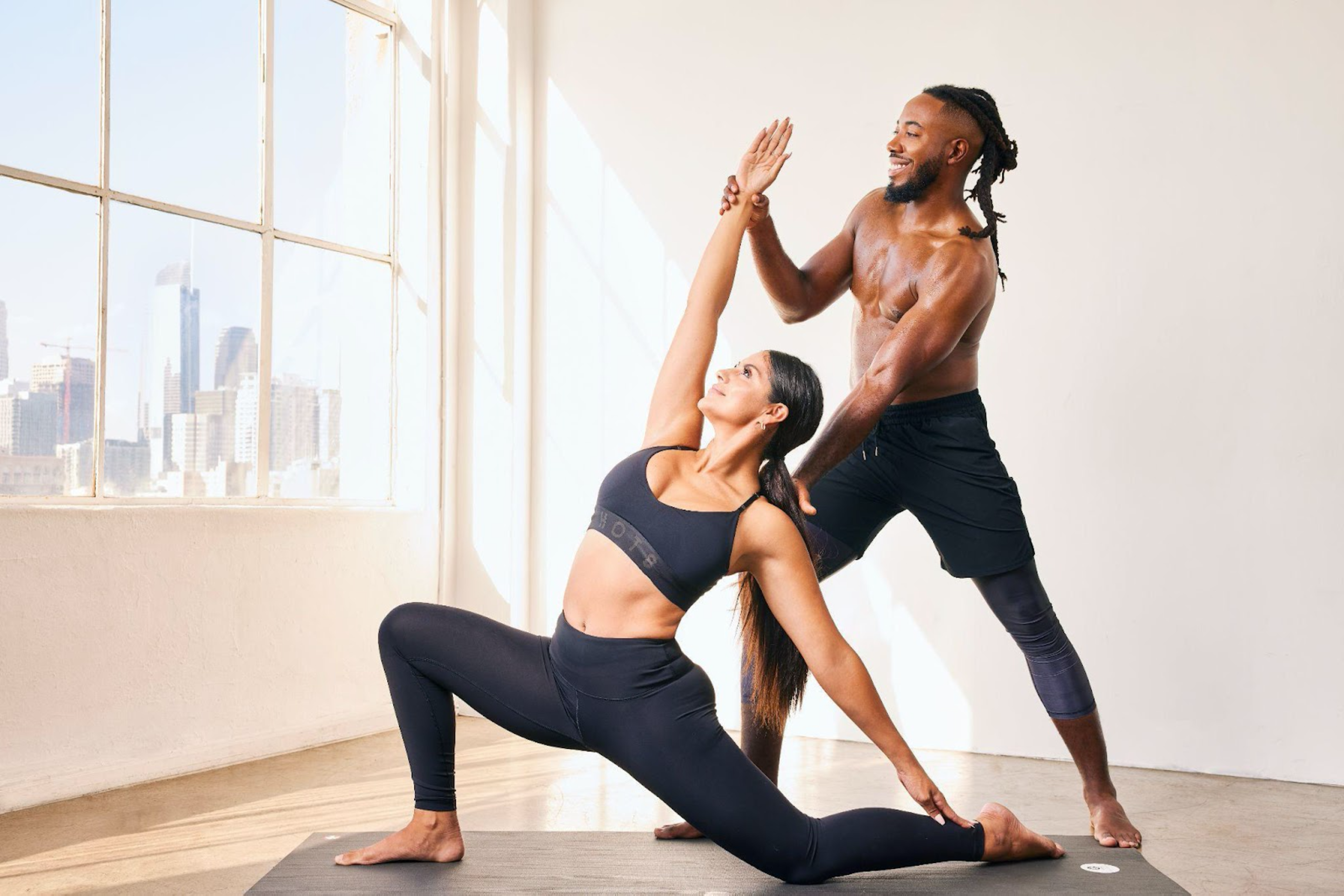 A woman in black workout clothes stretches in a yoga pose while a shirtless man in black shorts assists her in a bright room with city views.