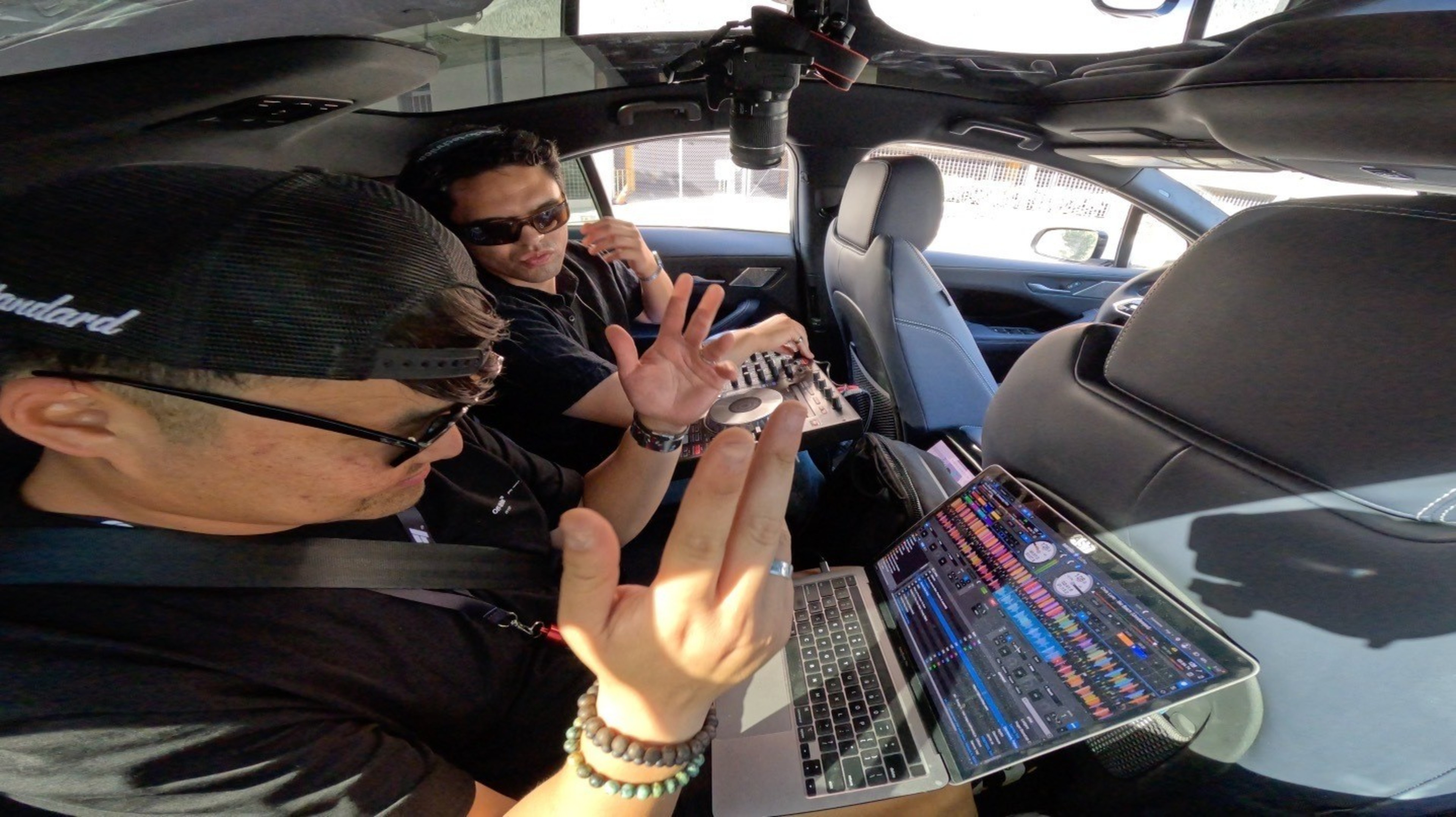 Two people in a car are using DJ equipment and a laptop. One wears a hat and gesticulates, while the other operates a mixer, both wearing sunglasses.