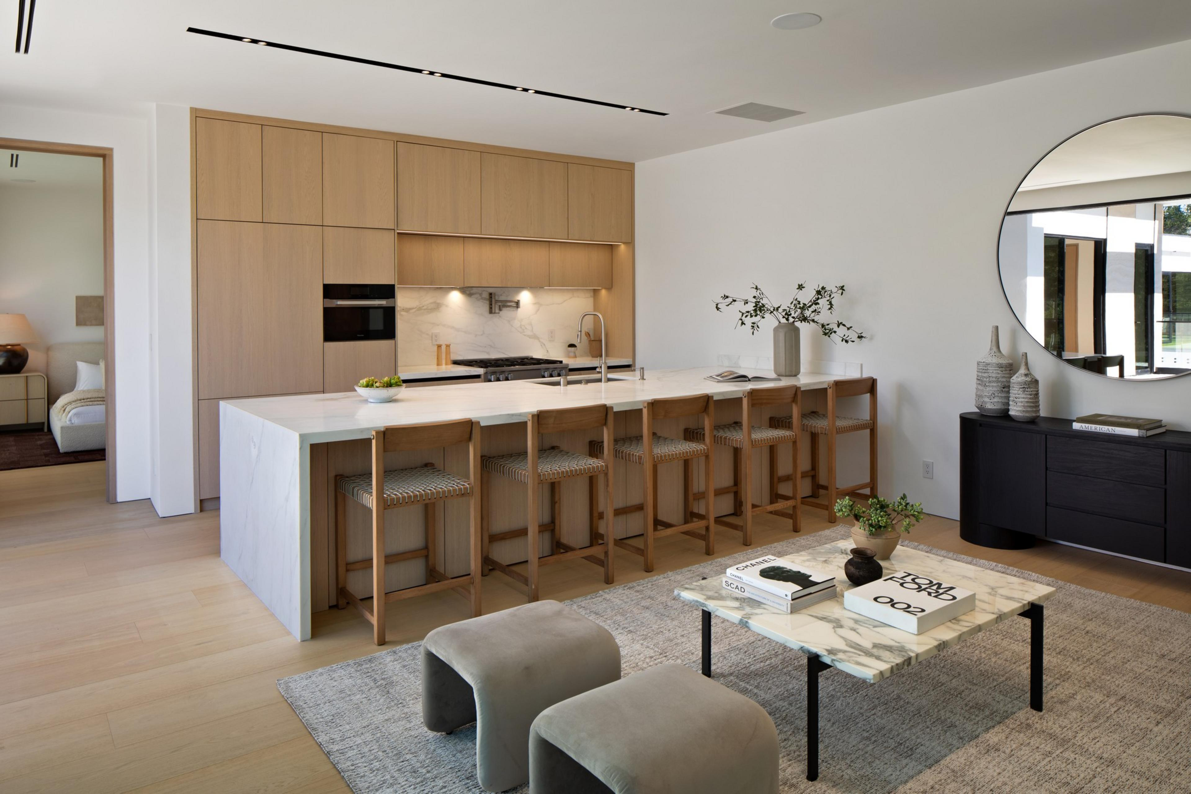 A modern kitchen with light wood cabinets, a white island with six chairs, a marble coffee table, two gray stools, and a round wall mirror.