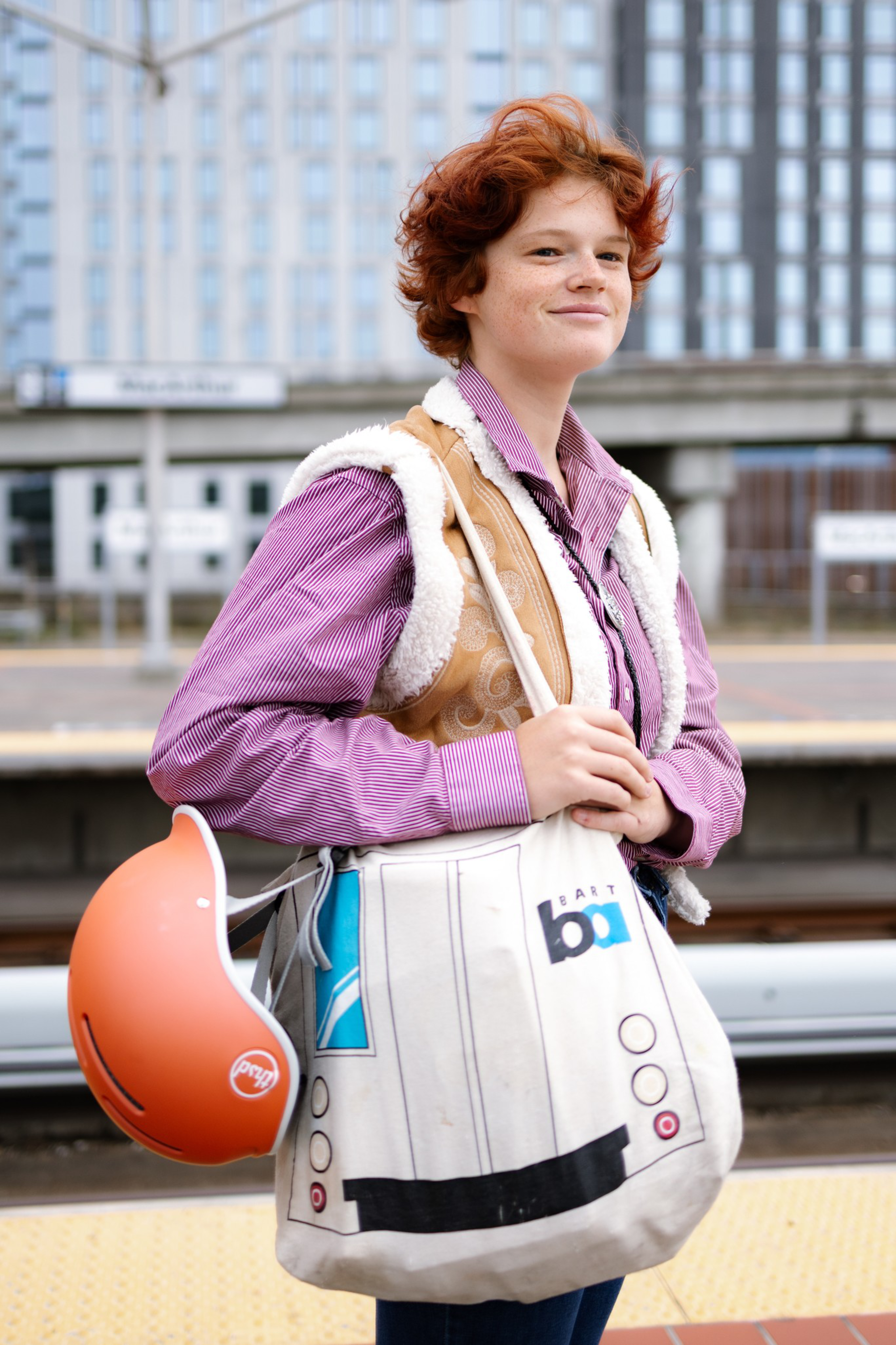 A person with short curly red hair wears a purple striped shirt and tan vest, holding a white BART tote bag and an orange helmet.