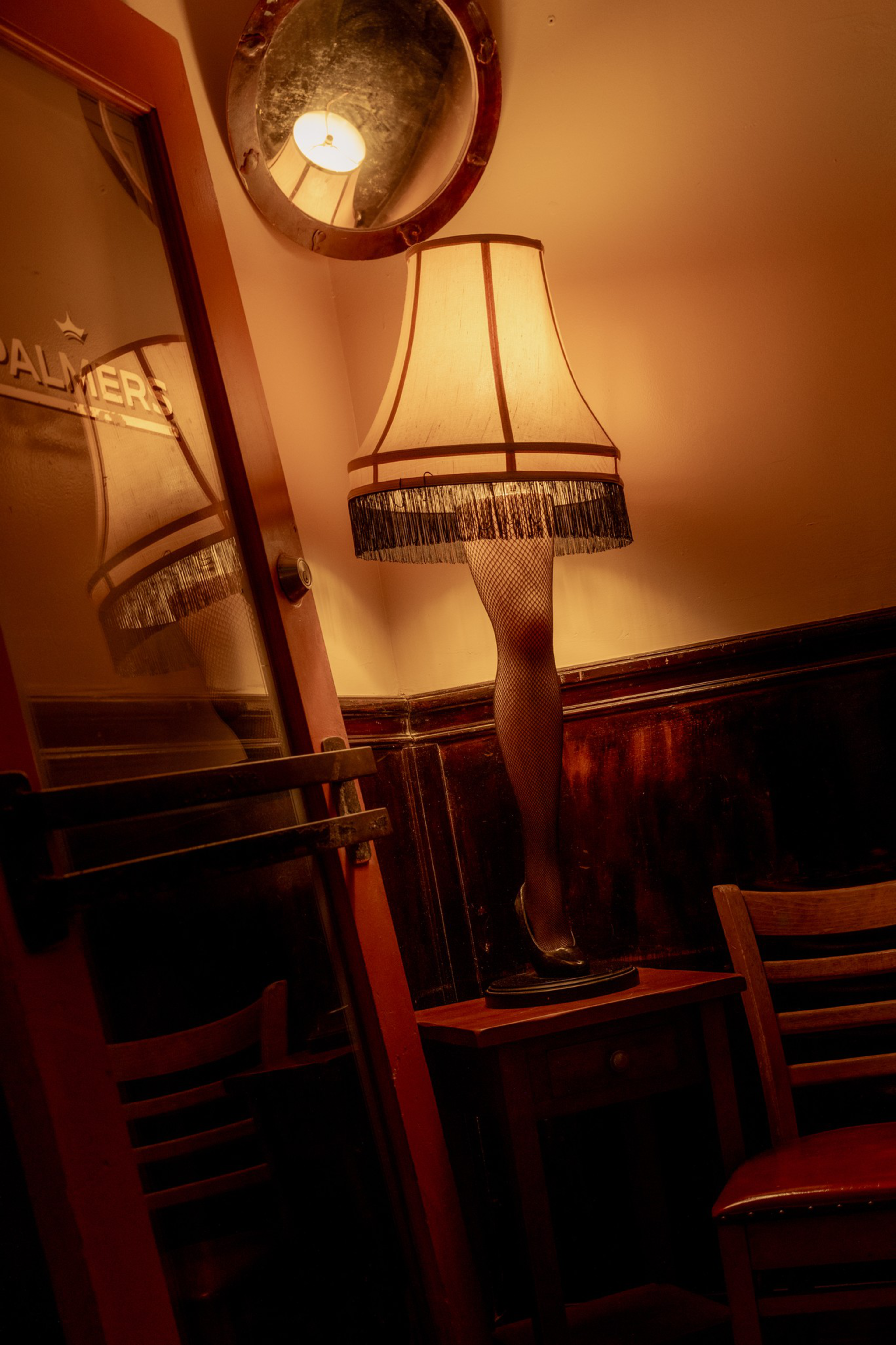 A lamp with a lampshade shaped like a fishnet stocking-covered leg with a high heel sits on a small table near wooden chairs and a door with a mirror.