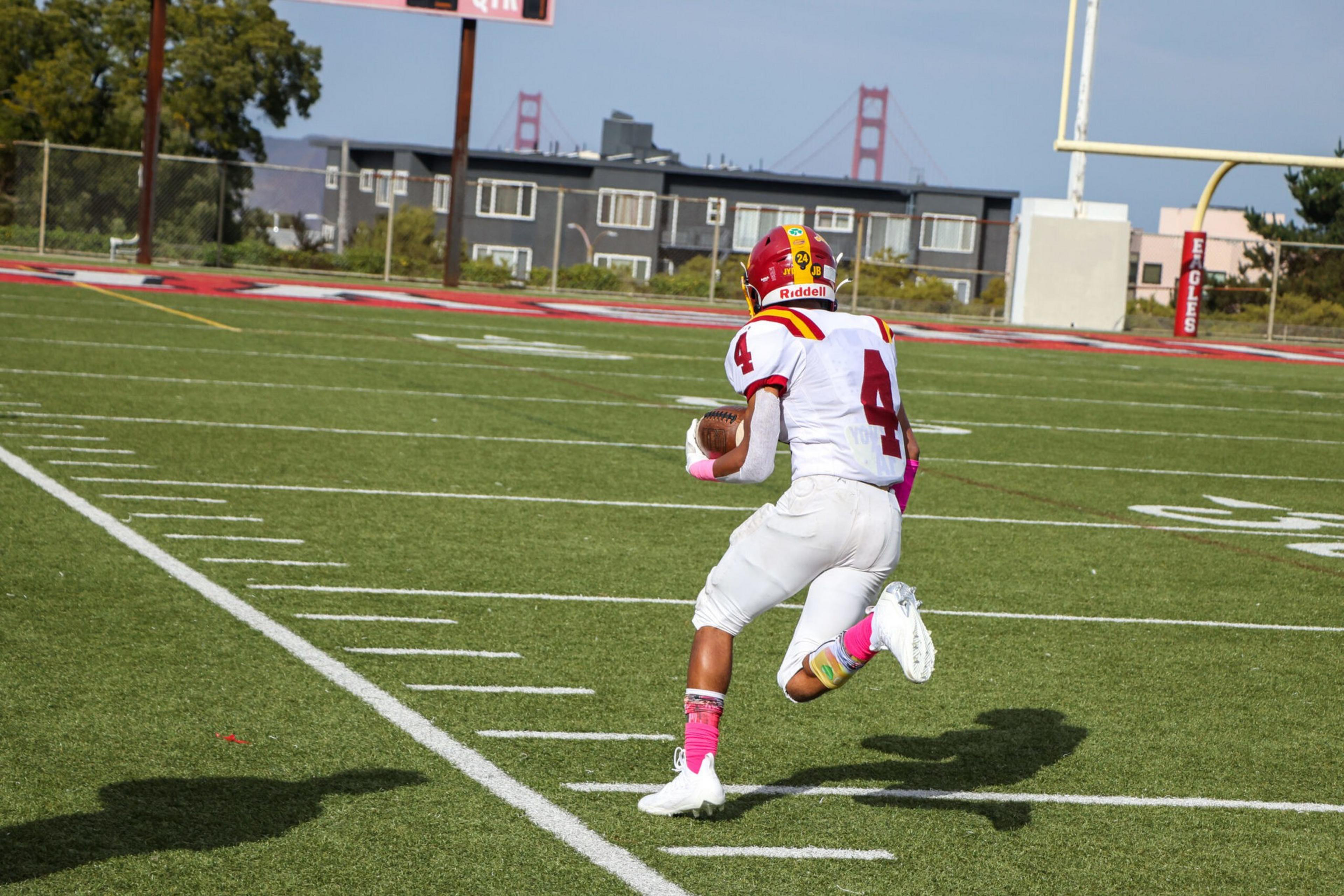 A football player wearing a white uniform with red and yellow accents and the number 4 runs on a green field holding a football.