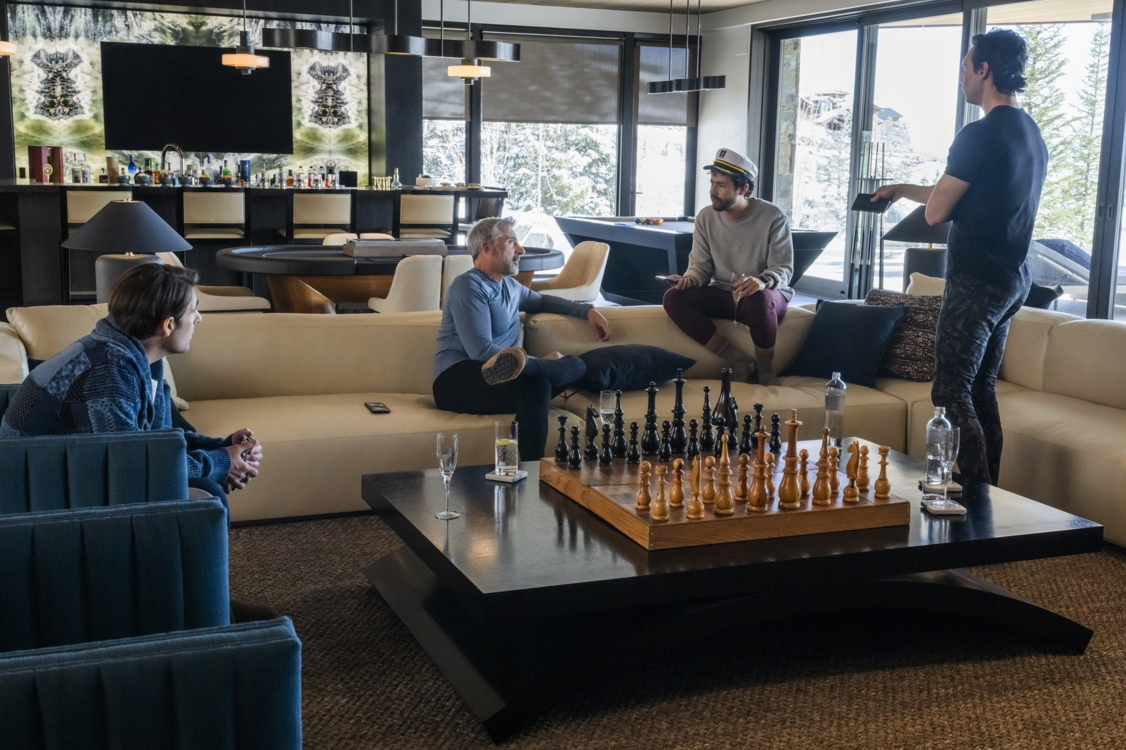Four people are gathered in a modern living room with large windows. A chess set is on the table, and bottles are in the background. It's a relaxed setting.