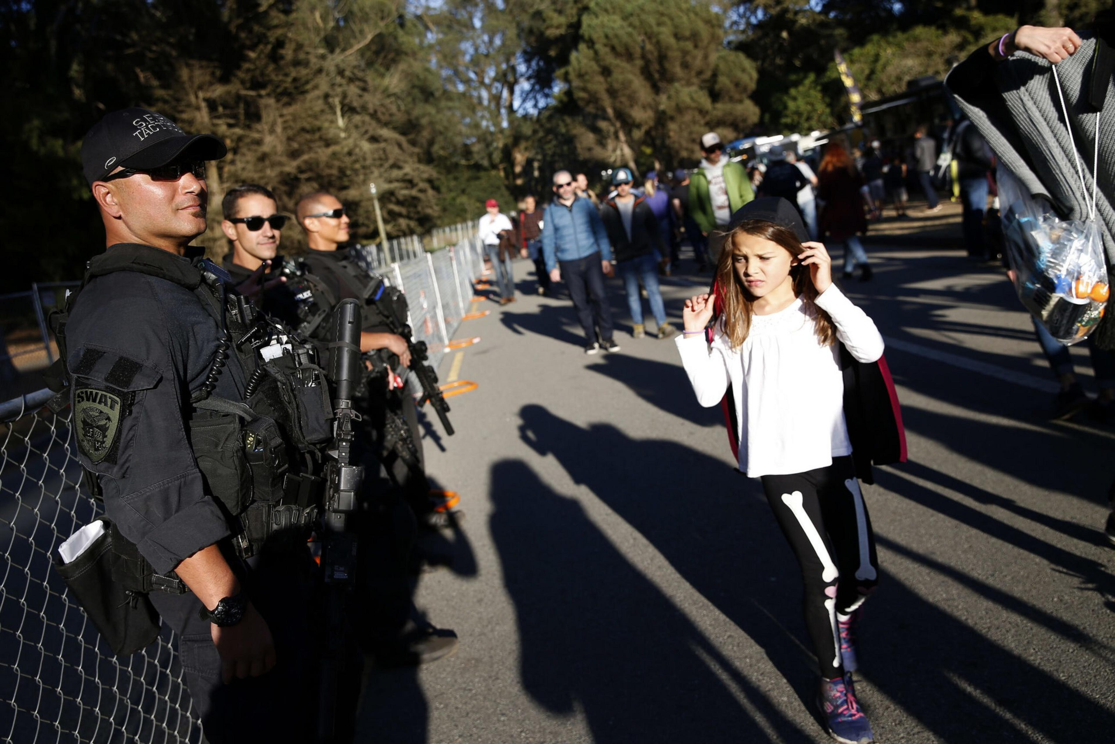 SWAT officers stand guard along a fence while a girl in a skeleton costume walks past under sunlight, with other people in the background.