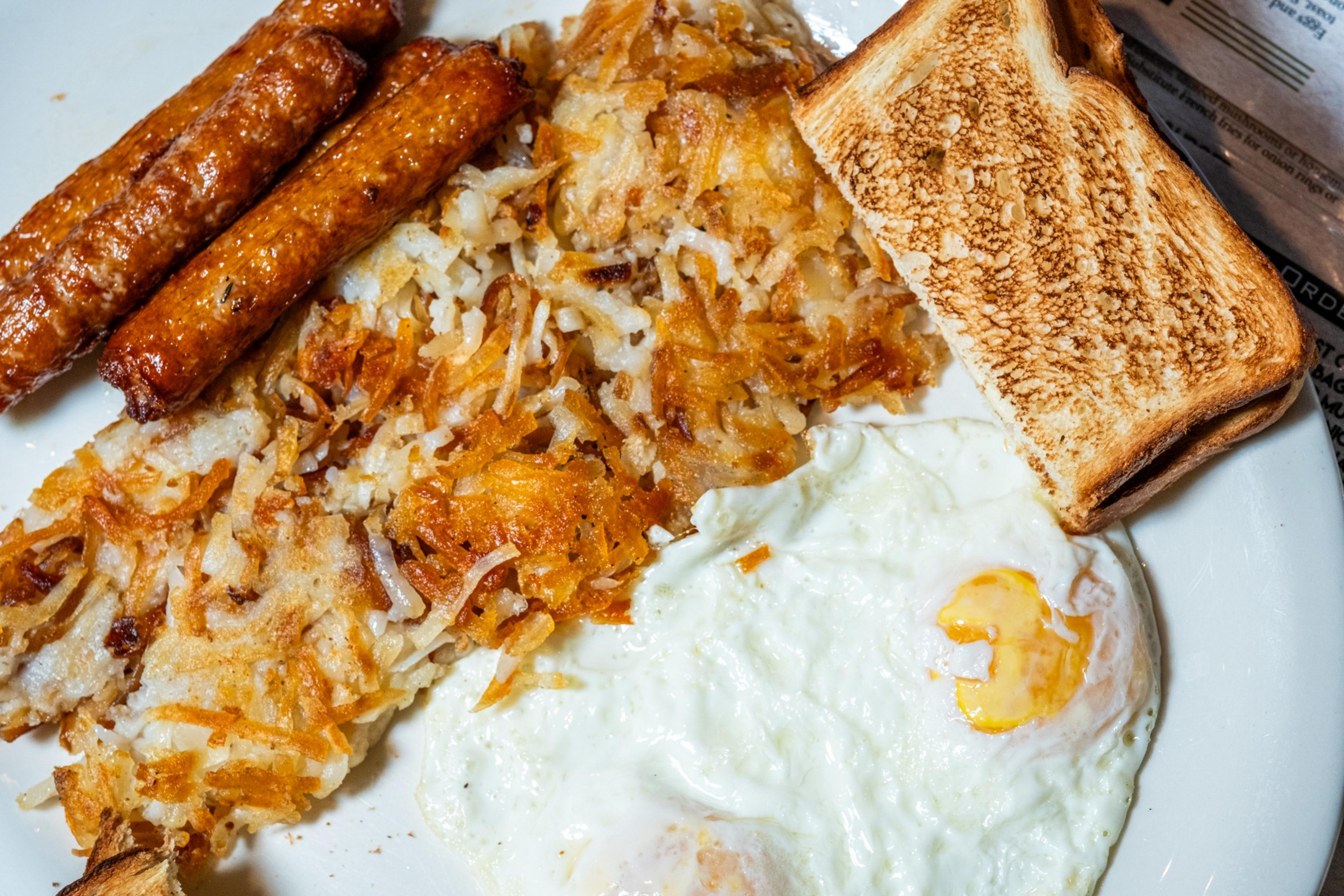 The image shows a breakfast plate with three sausages, crispy hash browns, two fried eggs, and a slice of toasted bread.