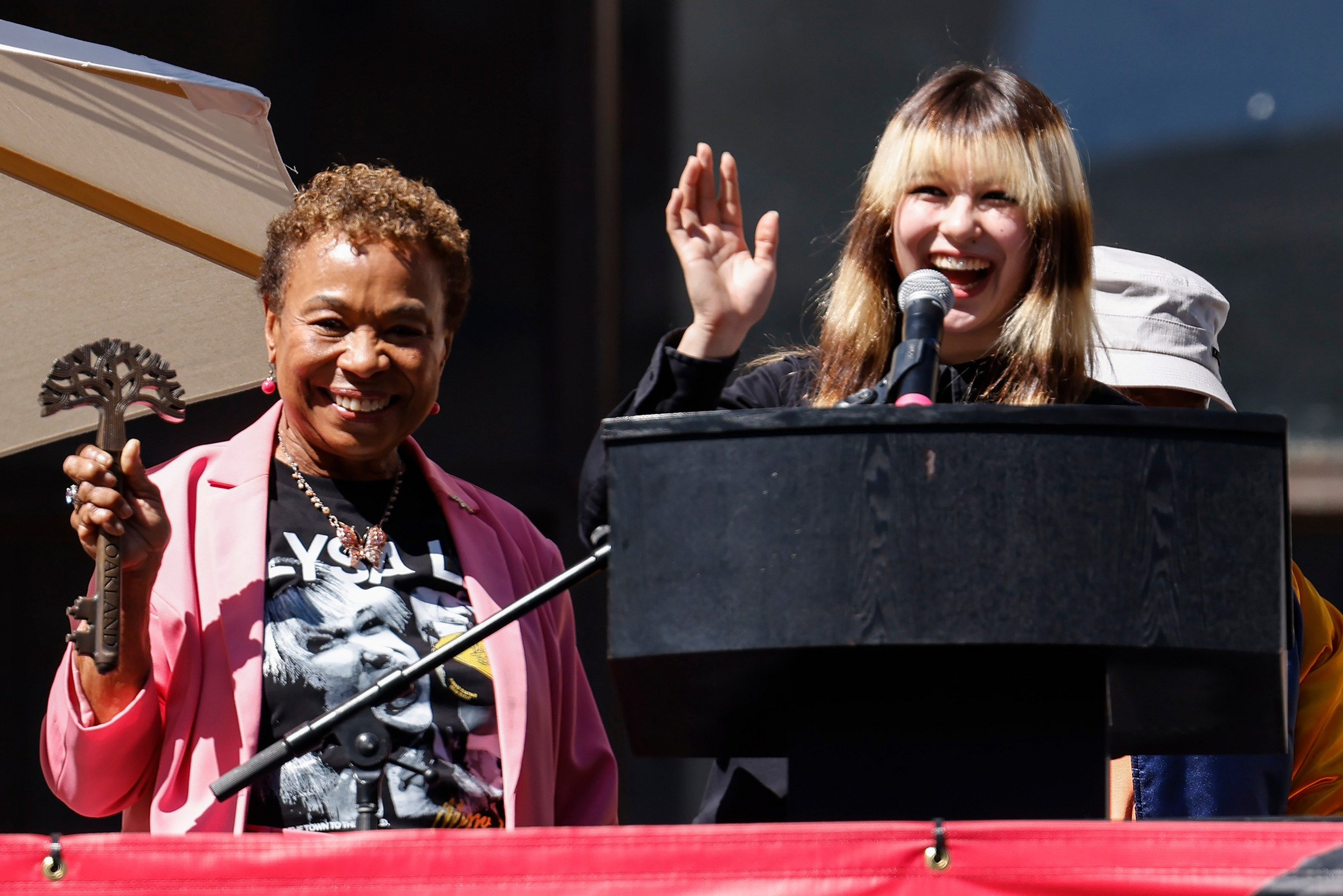 A smiling woman in a pink jacket holds a large ornamental key, while a woman at a podium with a microphone waves and smiles.