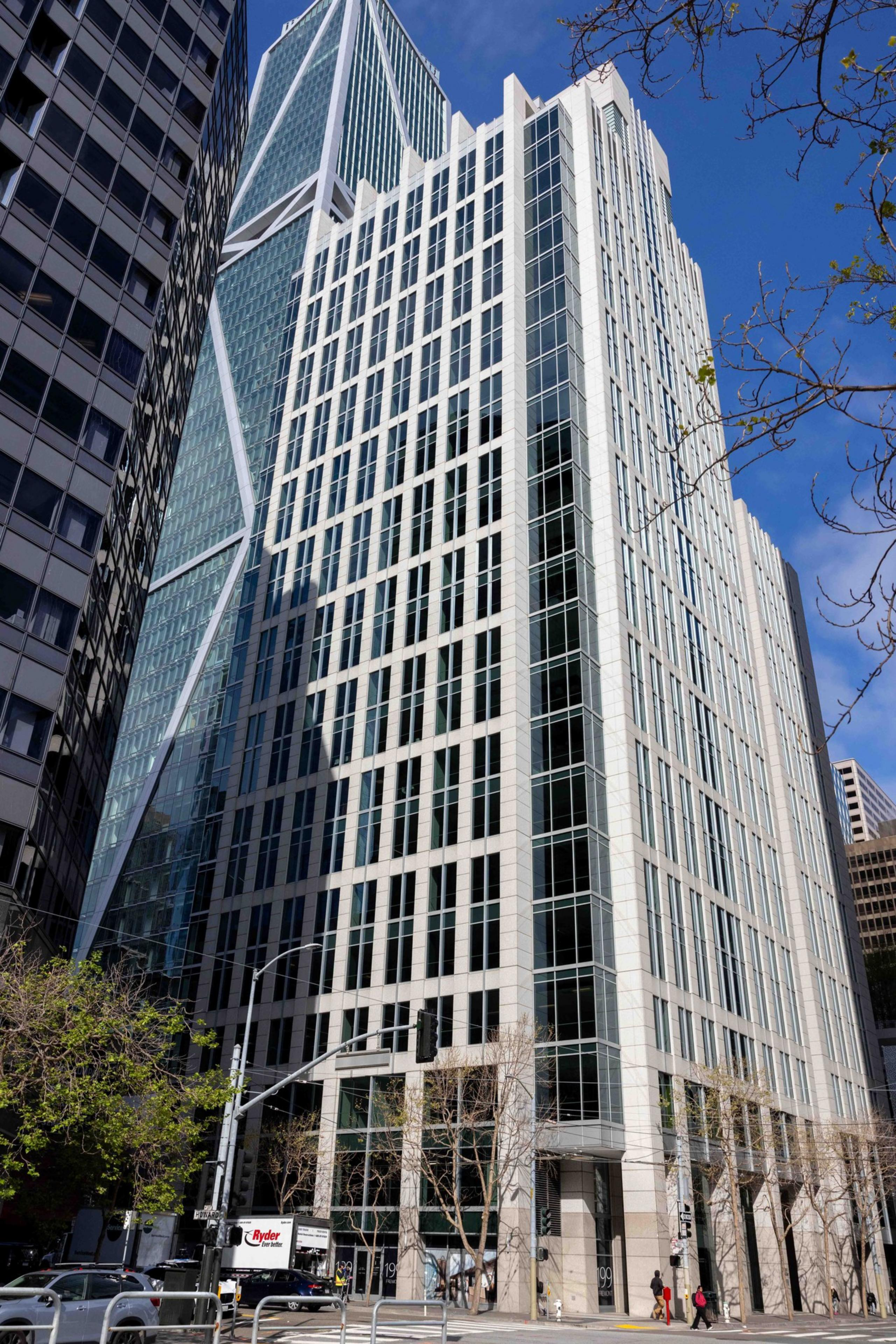 A tall, modern skyscraper with a sleek glass and white exterior stands against a blue sky. Nearby, leafless trees and a few pedestrians are visible.