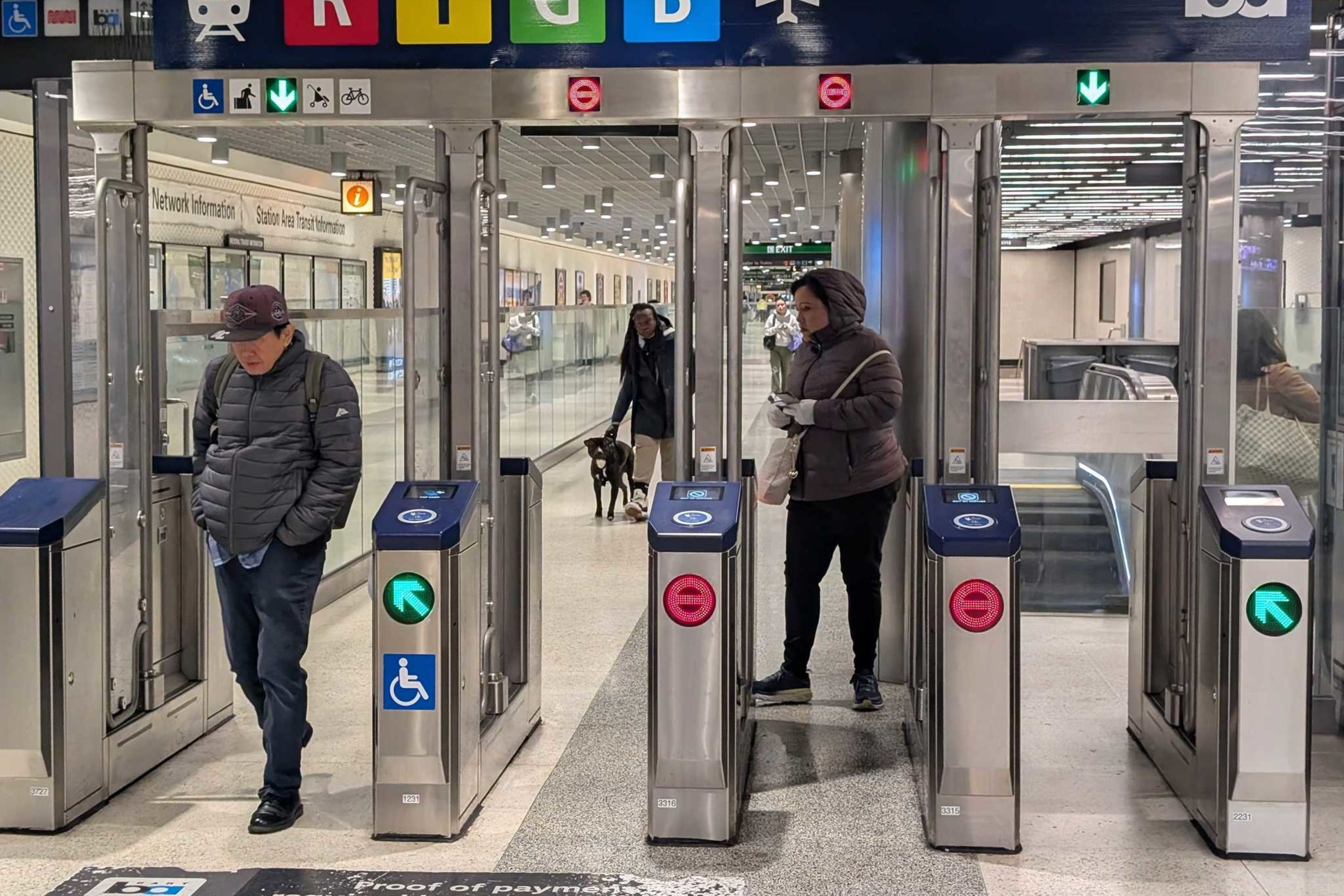 This image shows people passing through subway turnstiles at a station. The turnstiles have colored route indicators: Red, Yellow, Green, and Blue.