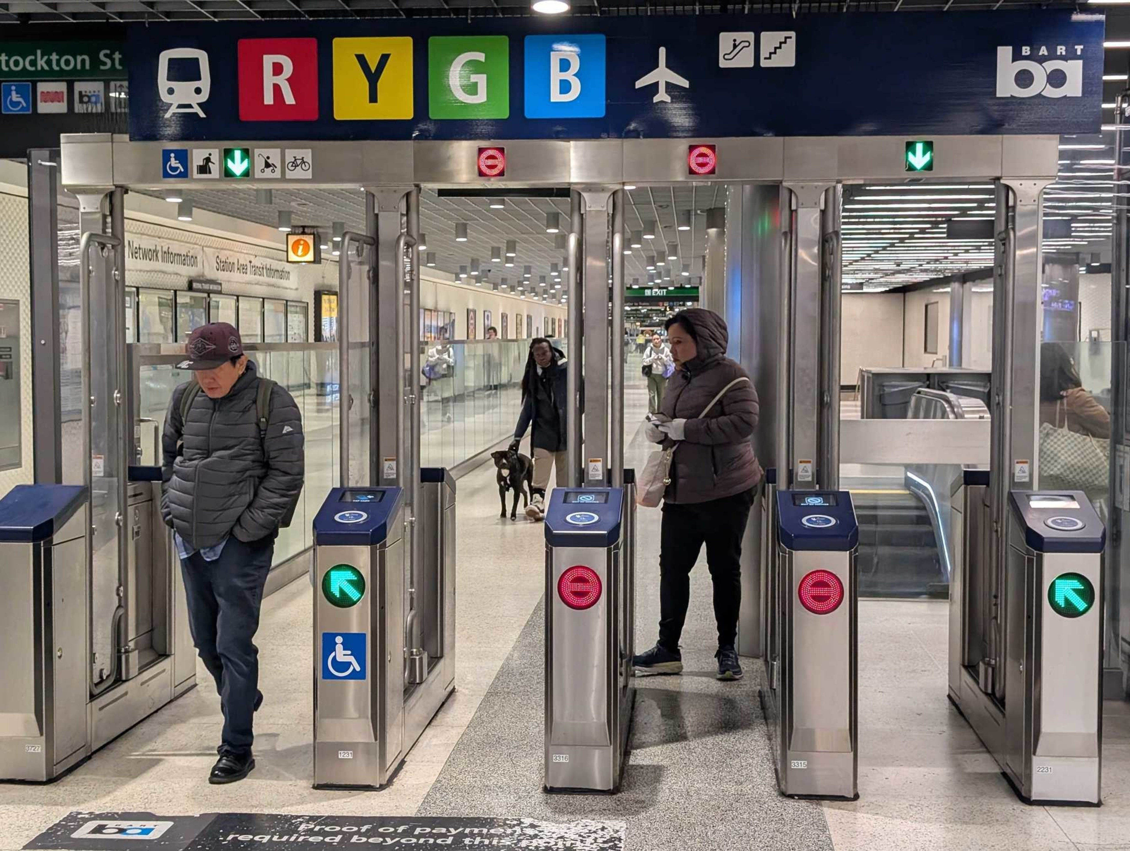 This image shows people passing through subway turnstiles at a station. The turnstiles have colored route indicators: Red, Yellow, Green, and Blue.