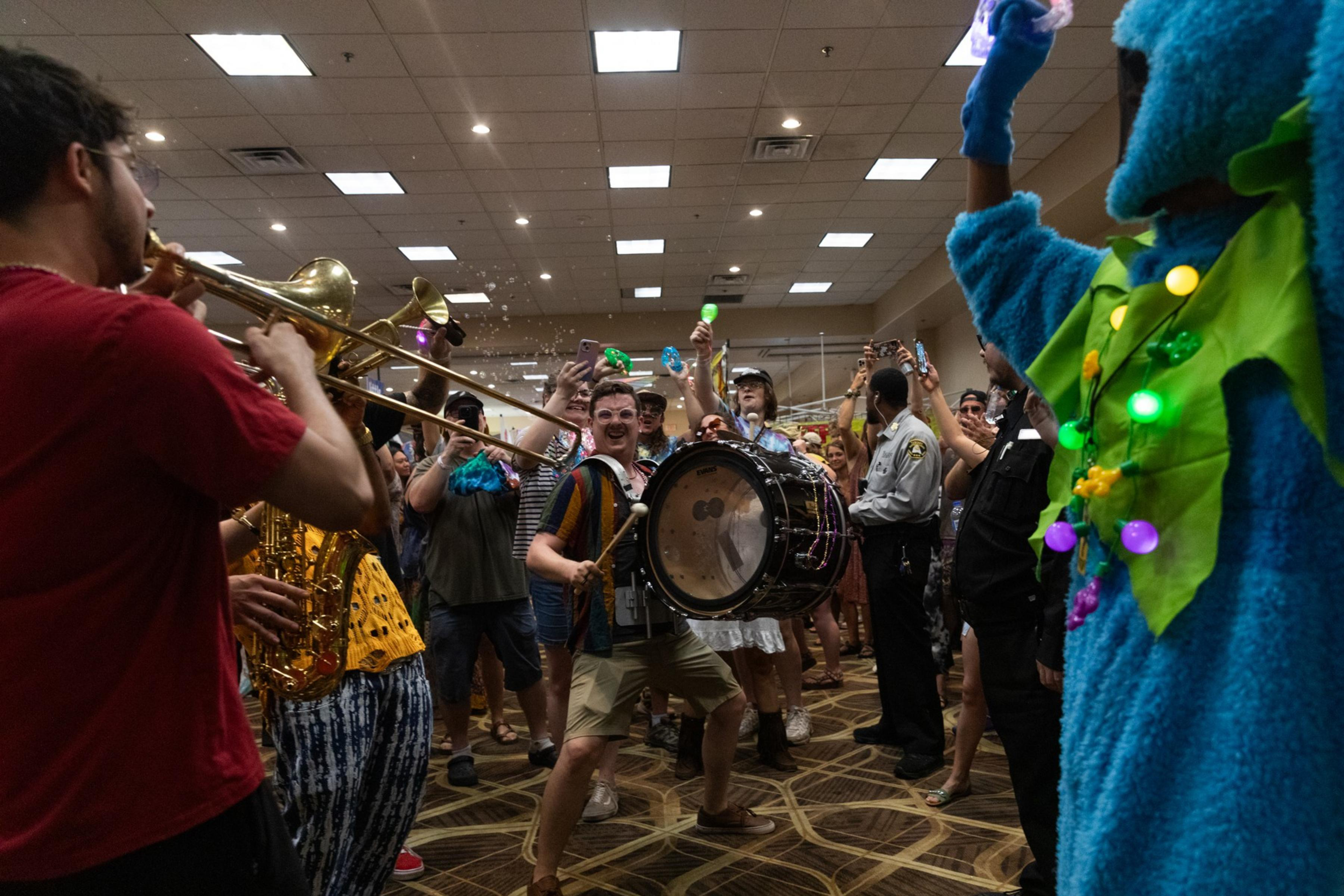 A lively indoor scene shows a costumed character in blue, illuminated by lights, dancing beside a band with brass instruments, and a cheering, colorful crowd.