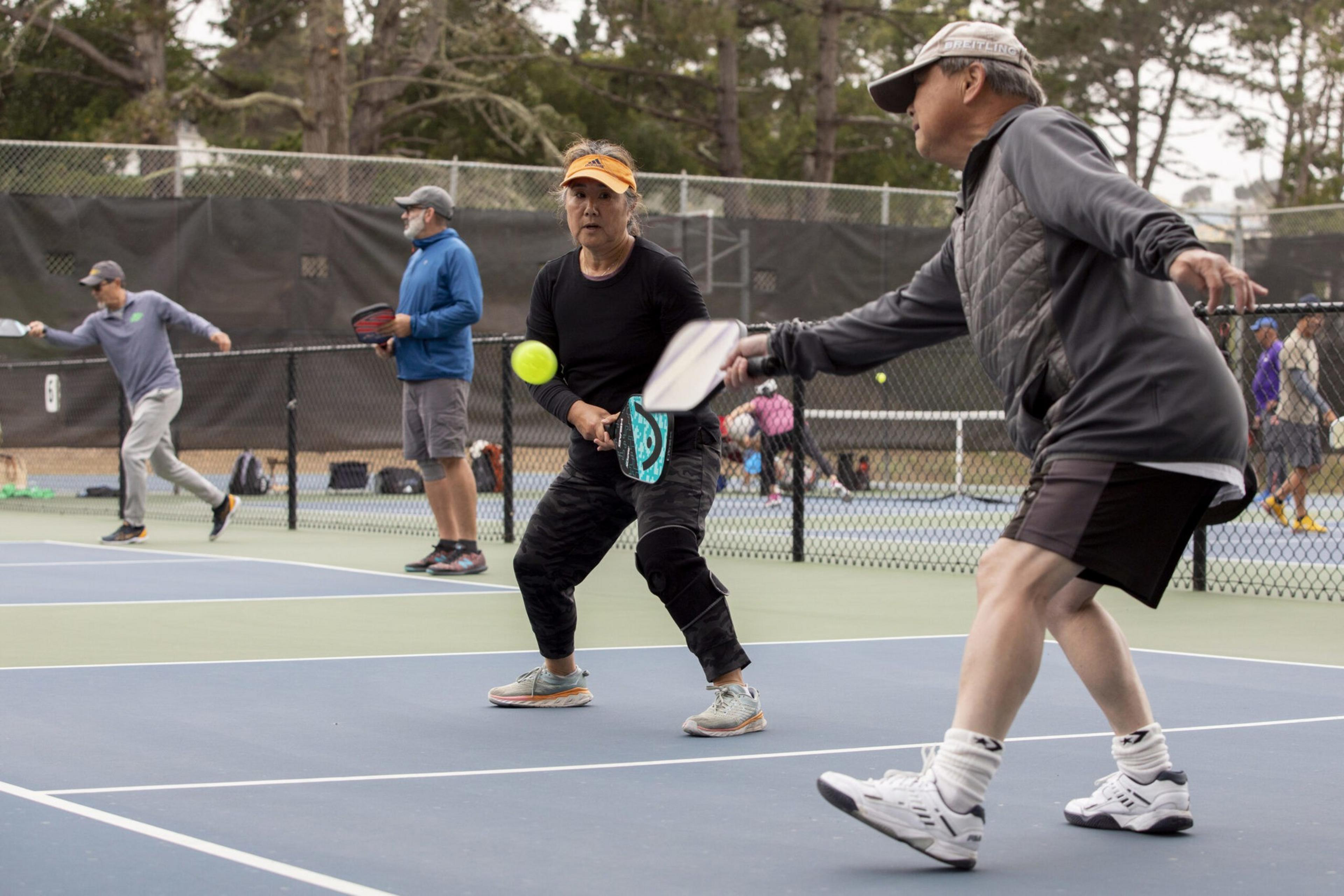 Two people are playing pickleball on an outdoor court, with one about to hit a yellow ball using a paddle, while others play on adjacent courts.
