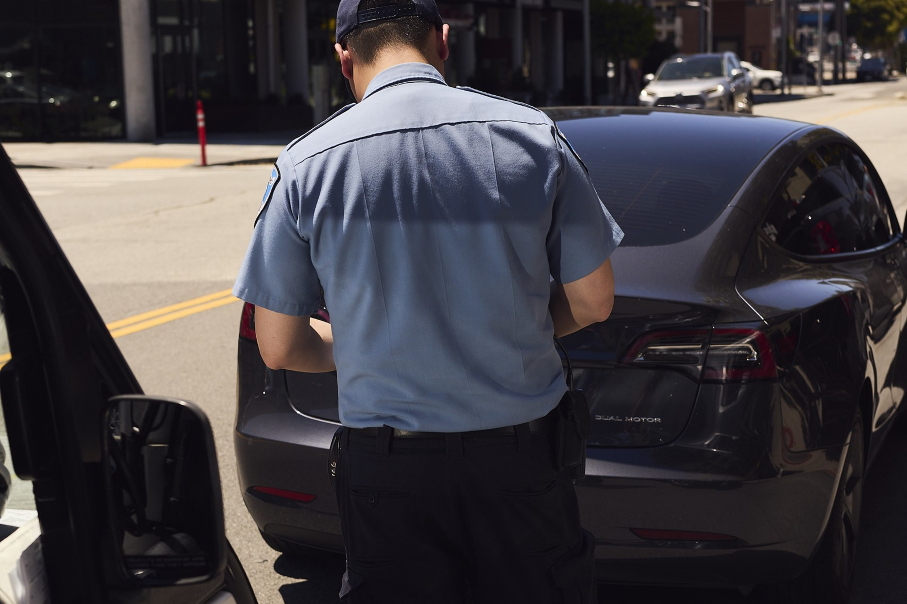 A uniformed officer stands on the street, writing a ticket beside a parked black car, with a building and other vehicles in the background.