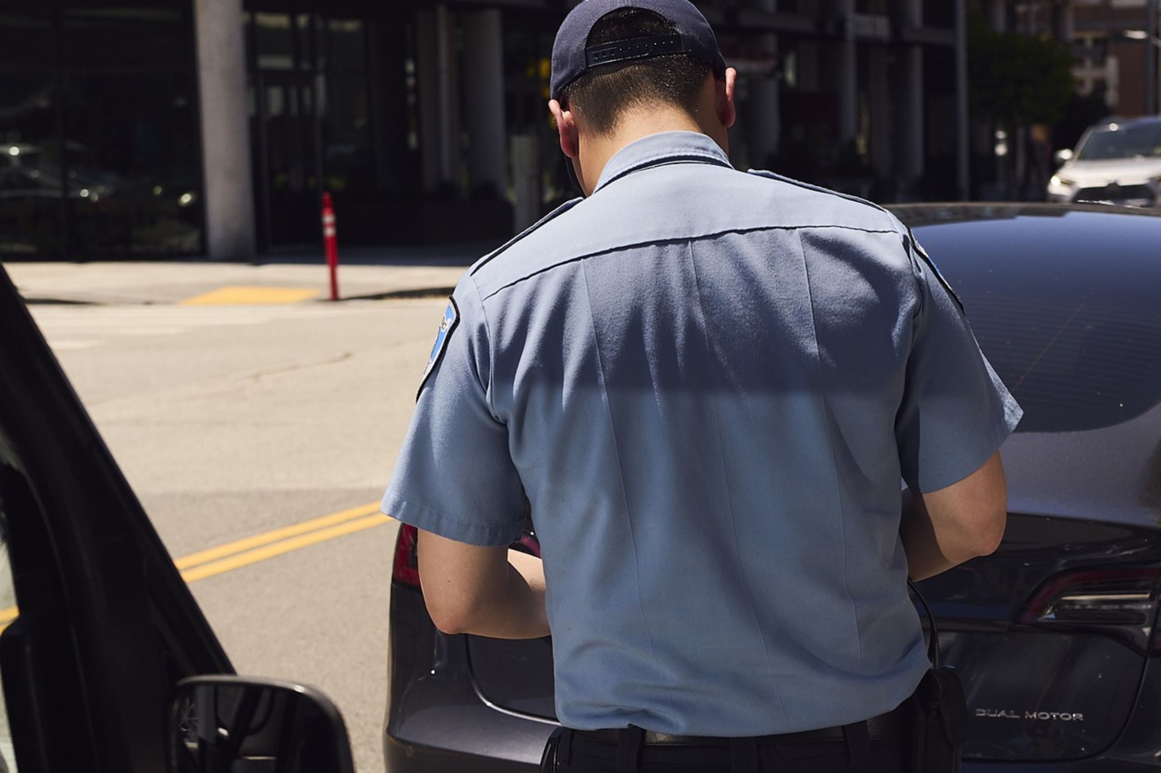 A uniformed officer stands on the street, writing a ticket beside a parked black car, with a building and other vehicles in the background.