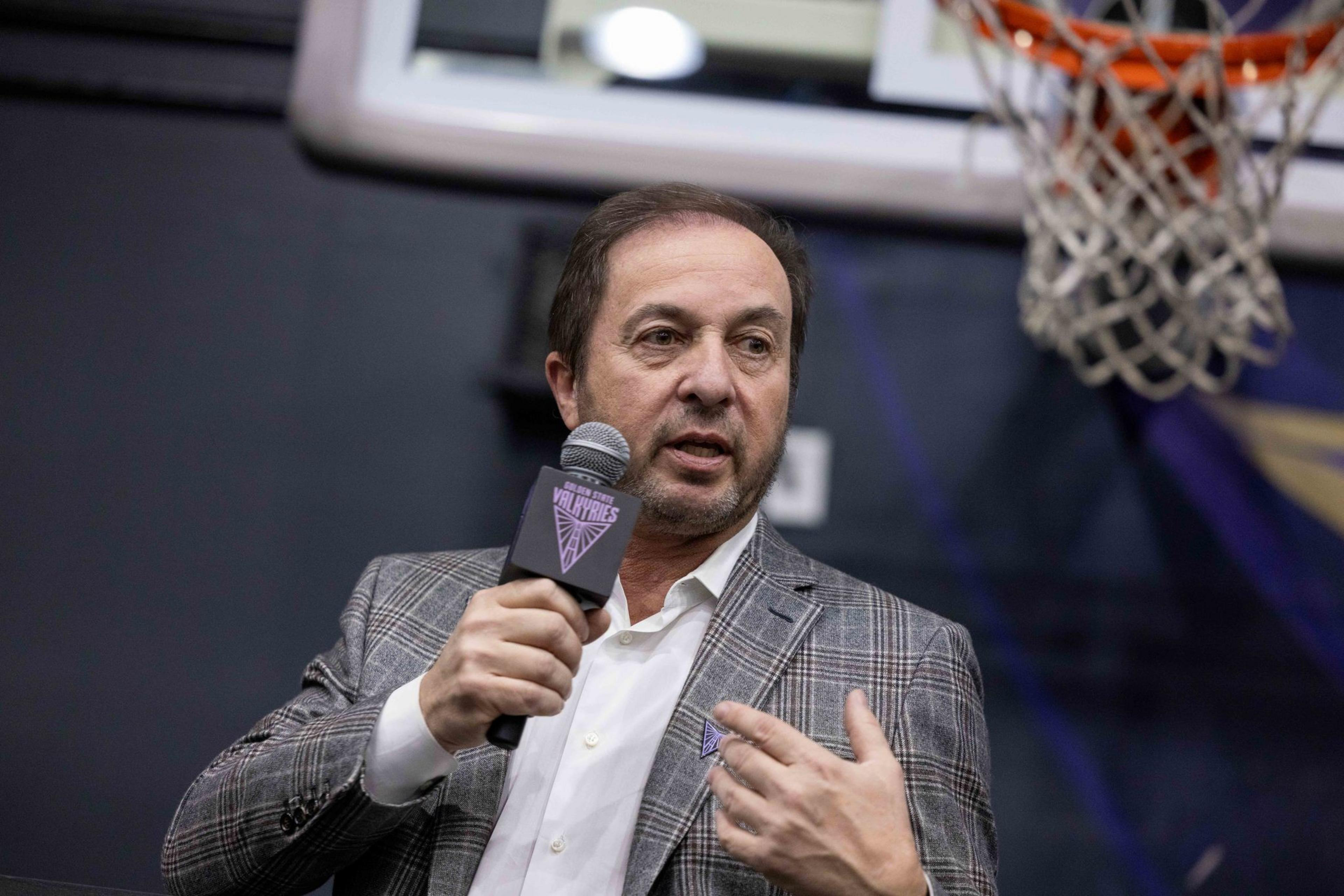 A man in a gray plaid suit speaks into a microphone with a sports logo. A basketball hoop is visible in the background.