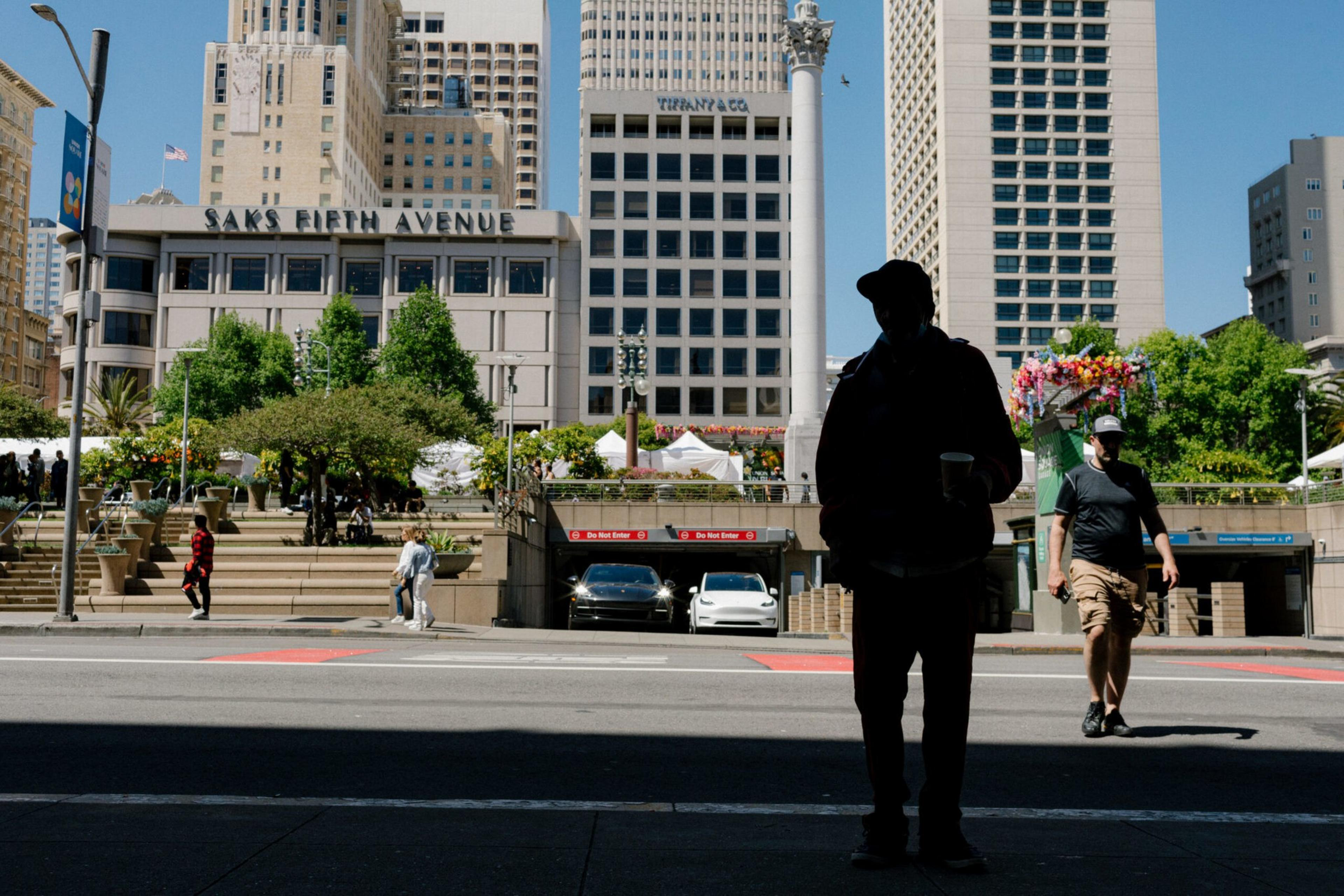 A city street with two people walking and one silhouetted, cars parked near stairs and several tall buildings labeled Saks Fifth Avenue and Tiffany & Co.