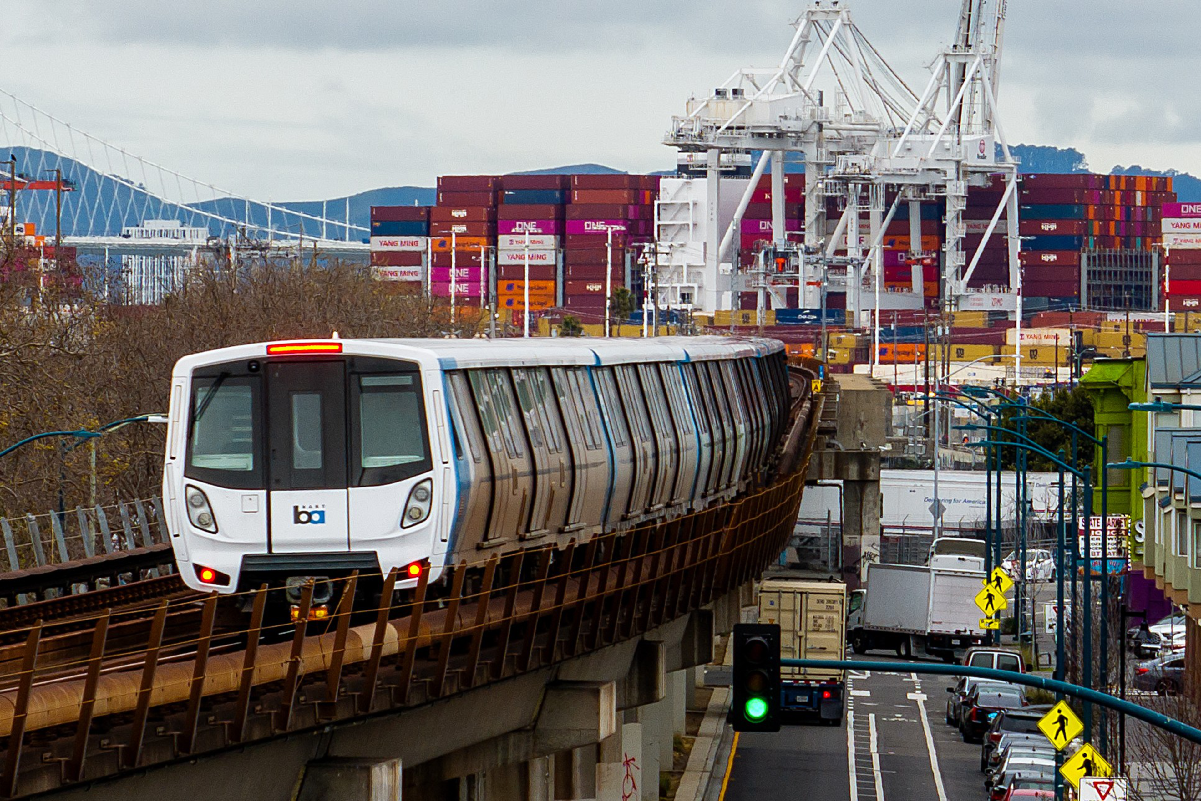 A train travels on an elevated track with a busy shipping port in the background. Shipping containers and cranes are visible under a cloudy sky.