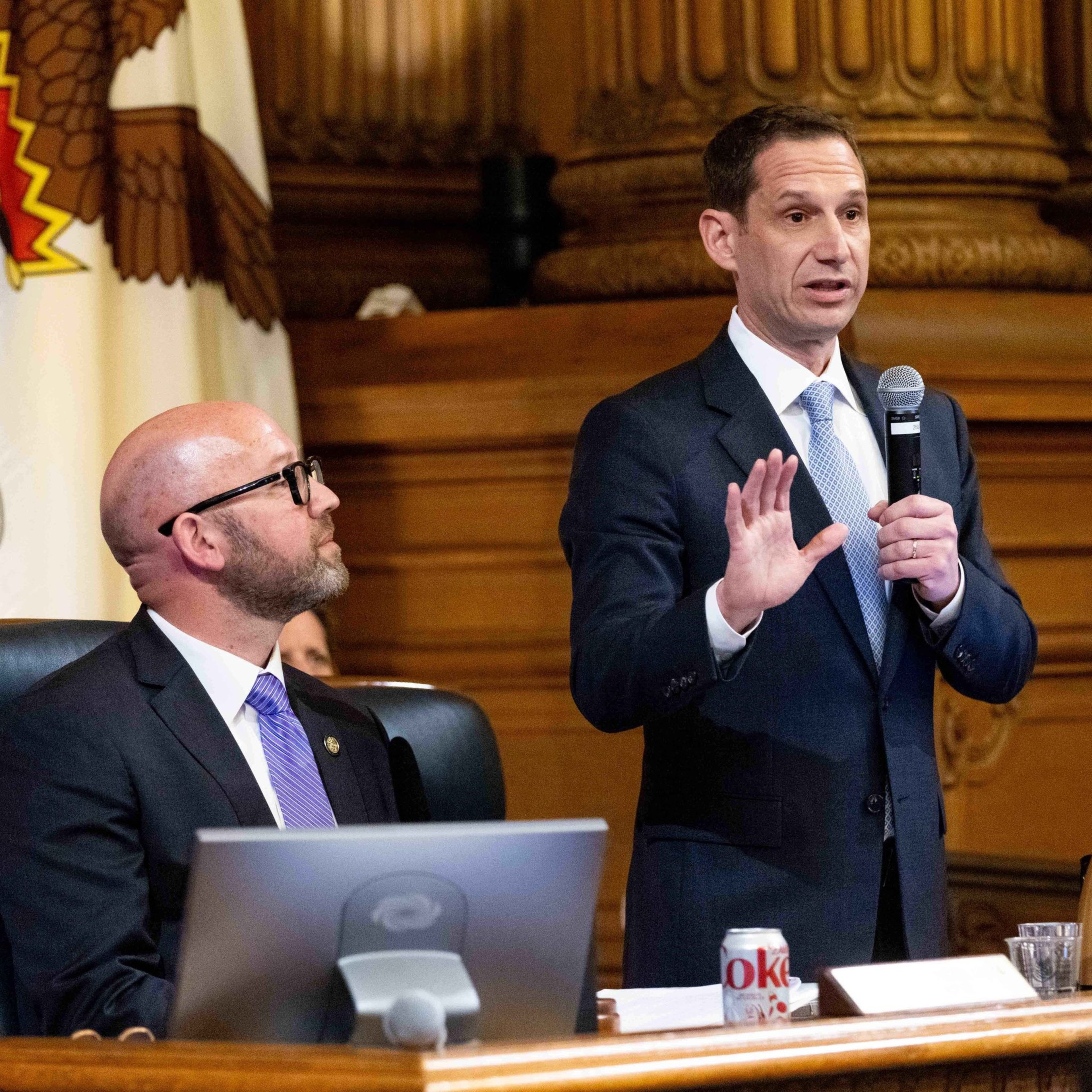 A man in a suit speaks into a microphone, gesturing, while another man in a suit sits at a desk with a laptop and Diet Coke in front of him. There's a US flag behind them.