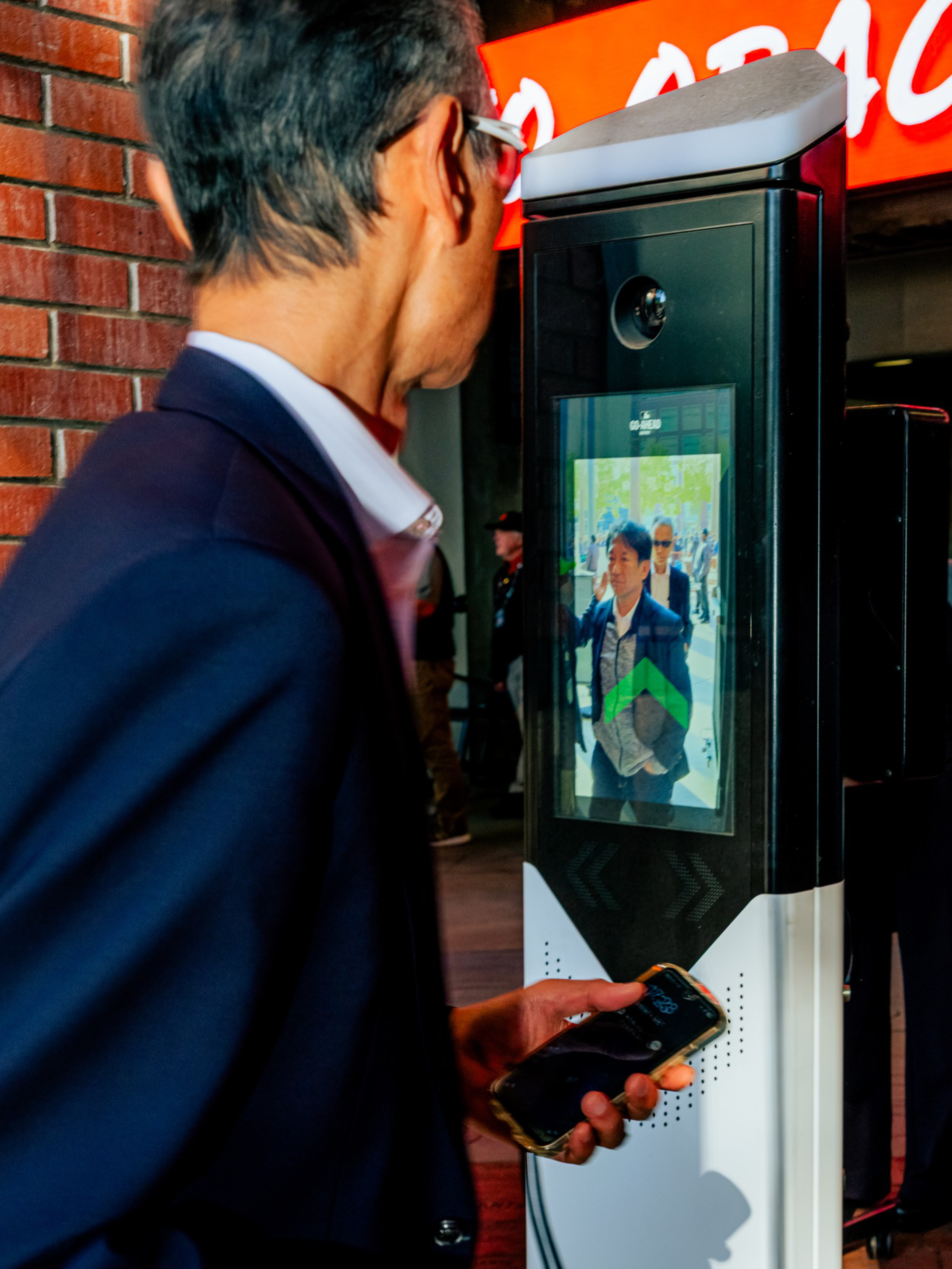 A man holding a phone looks at a tall digital kiosk displaying his face and a green arrow, standing near a red brick wall and a bright sign.