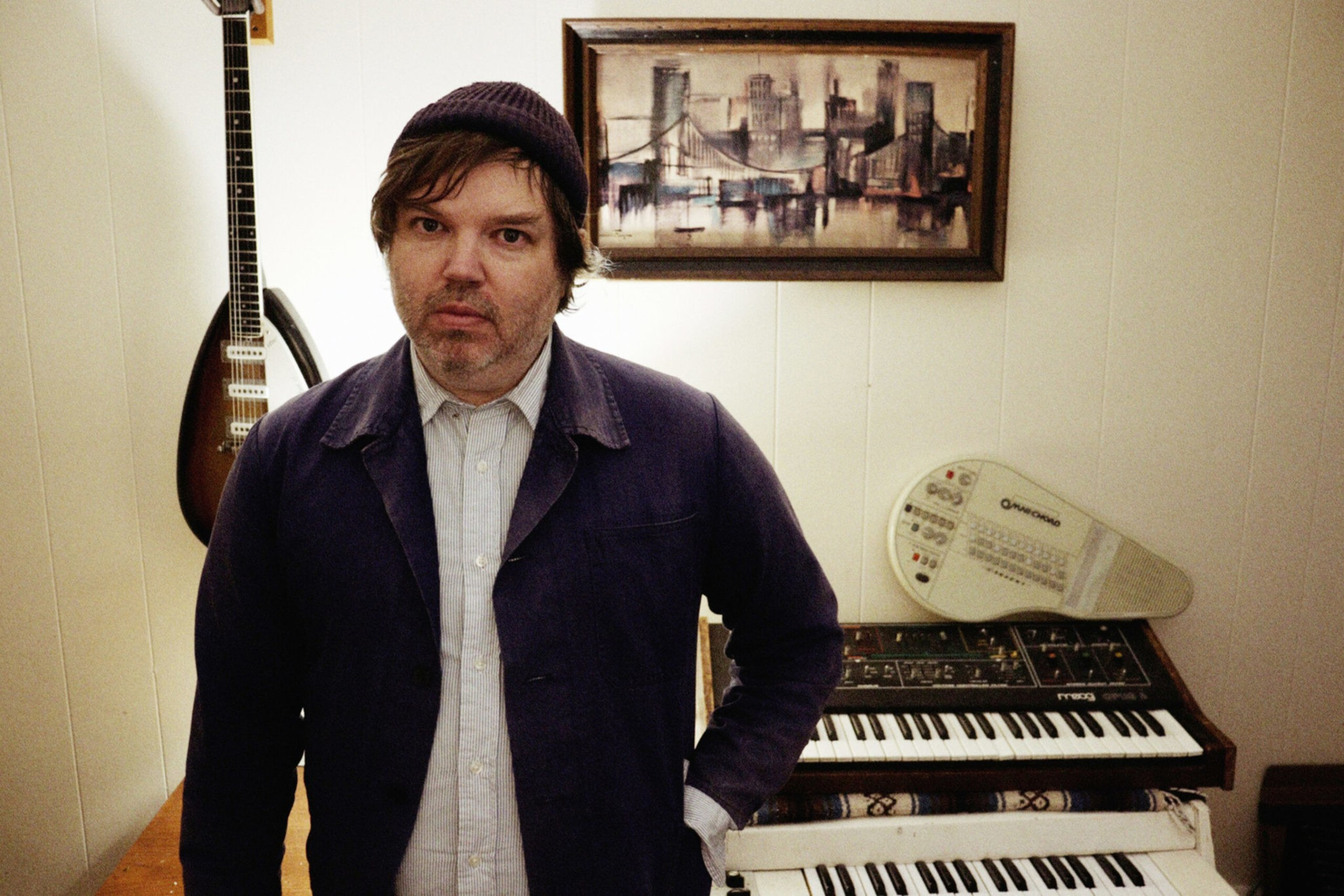 A man wearing a beanie and dark jacket stands in a room with a guitar, keyboard, and a framed cityscape painting on the wall behind him.