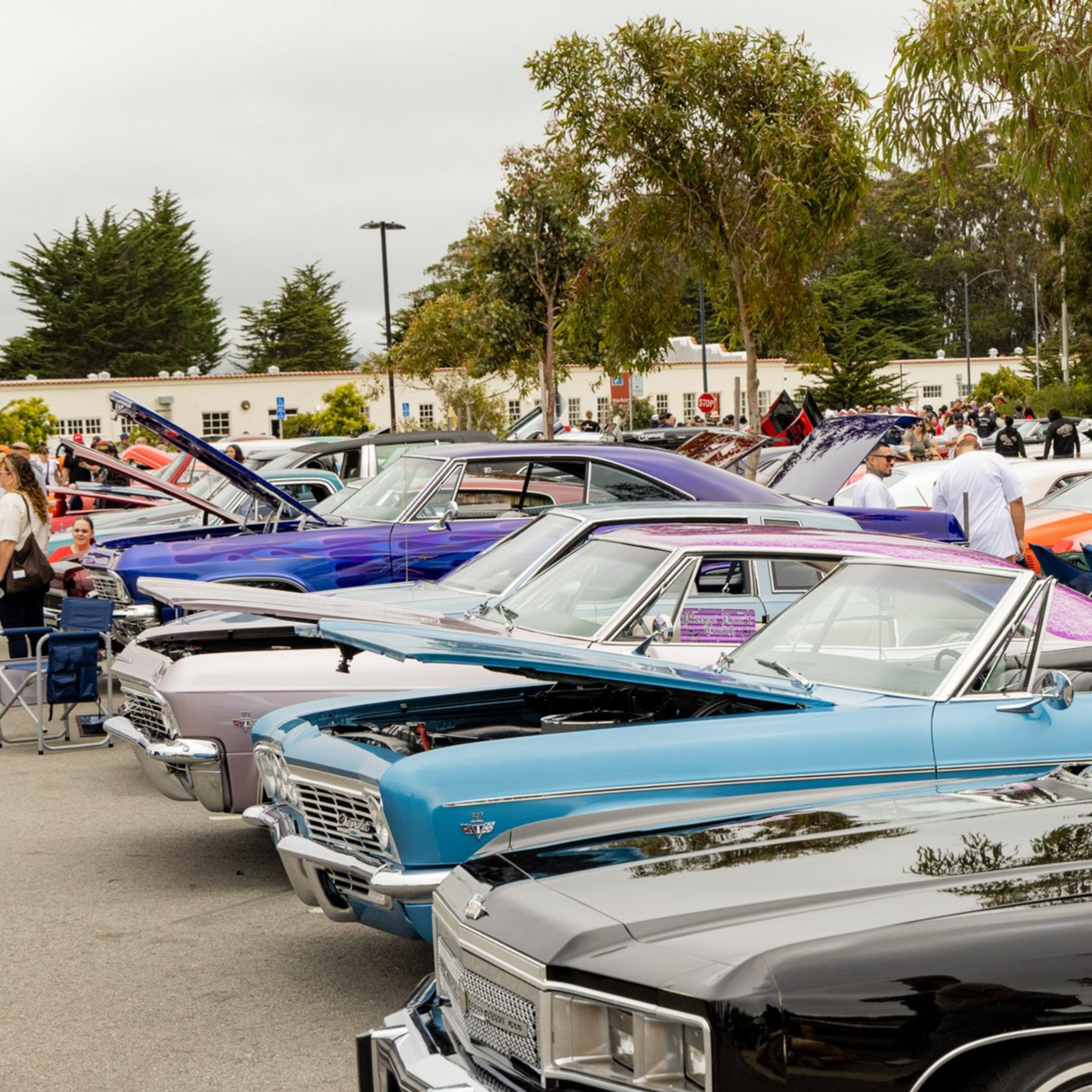 The image shows a line of classic cars with colorful exteriors parked at a car show. People are walking around, admiring the vehicles, and chatting.