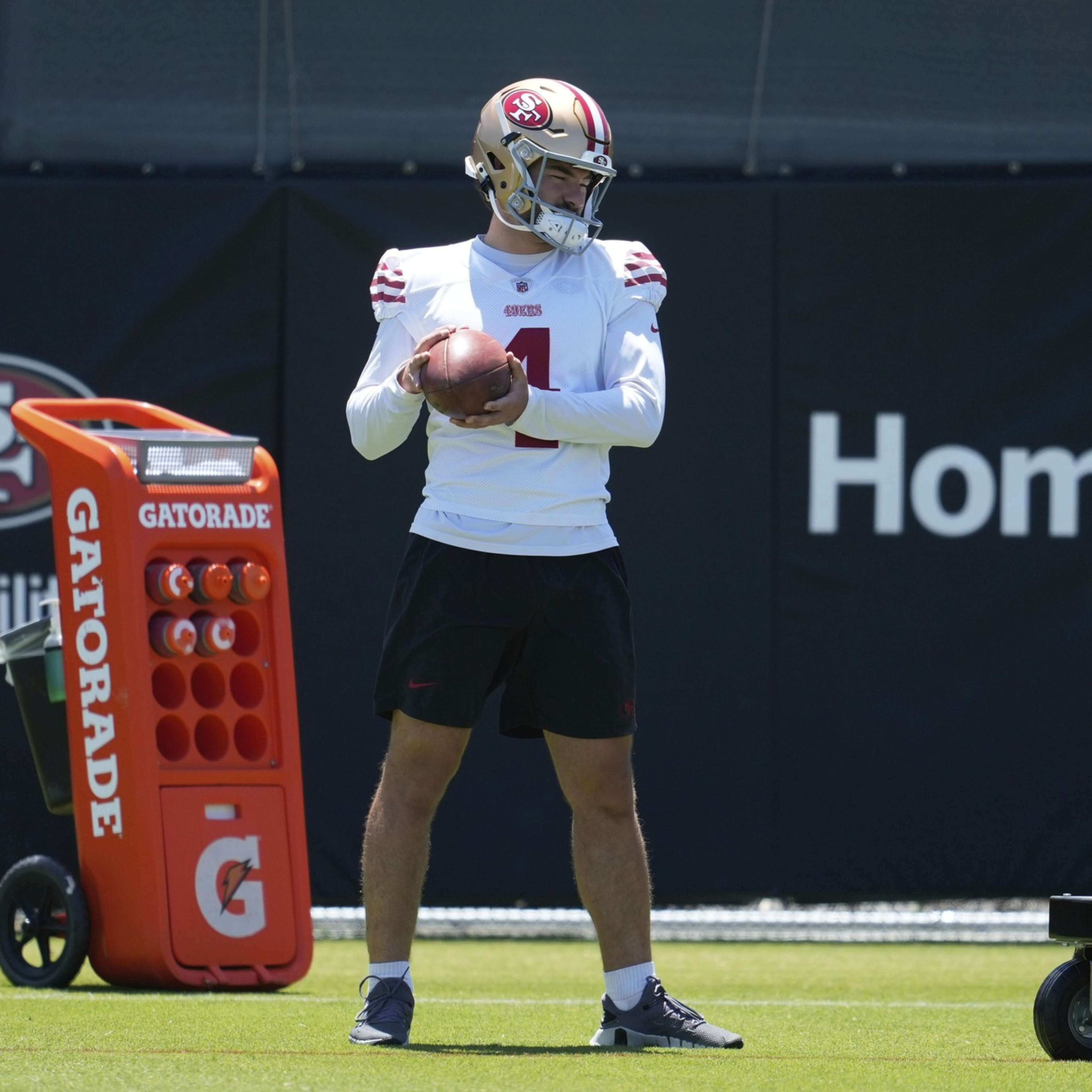 A football player wearing a San Francisco 49ers helmet and practice uniform holds a football, standing near an orange Gatorade cooler on the field.