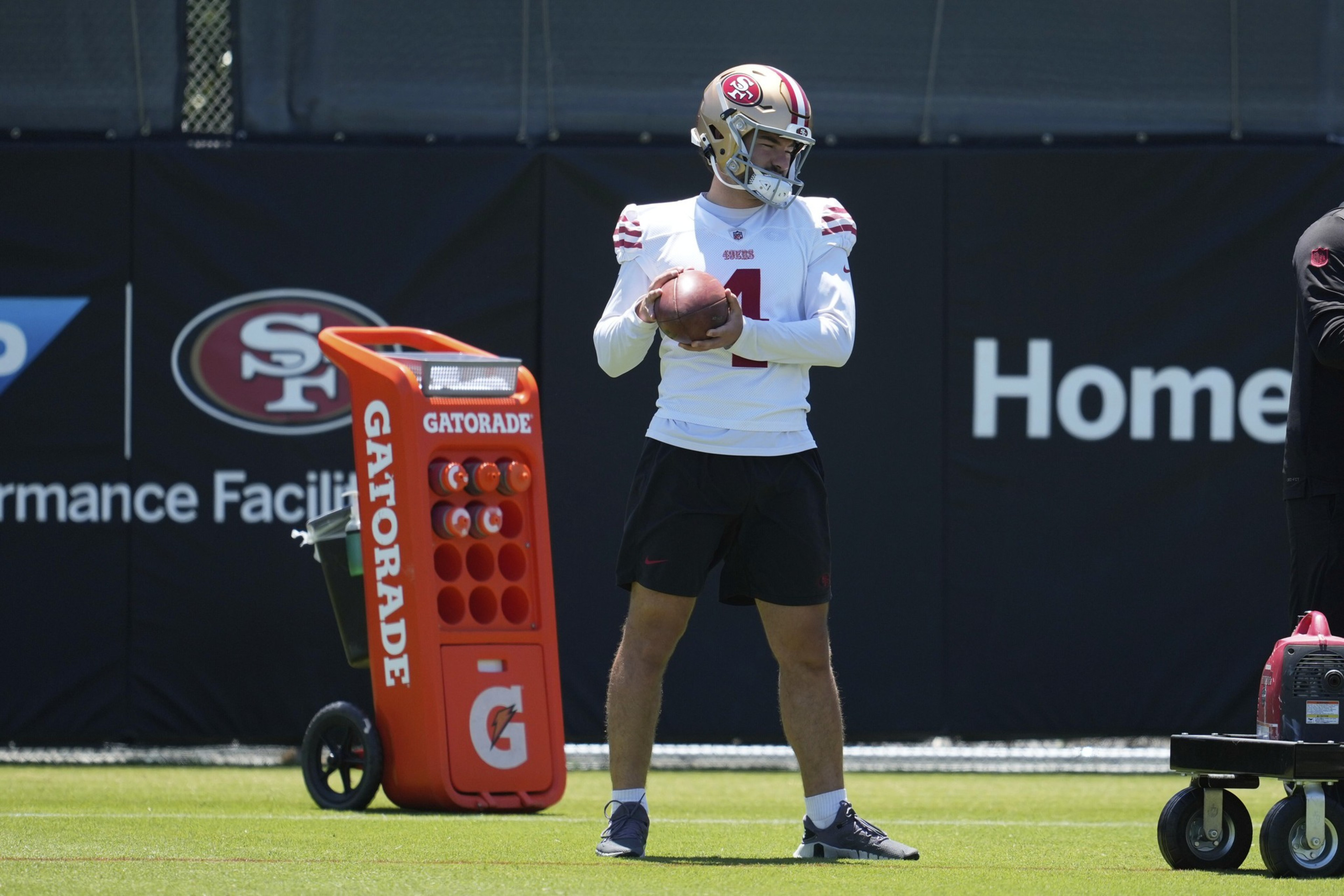 A football player wearing a San Francisco 49ers helmet and practice uniform holds a football, standing near an orange Gatorade cooler on the field.