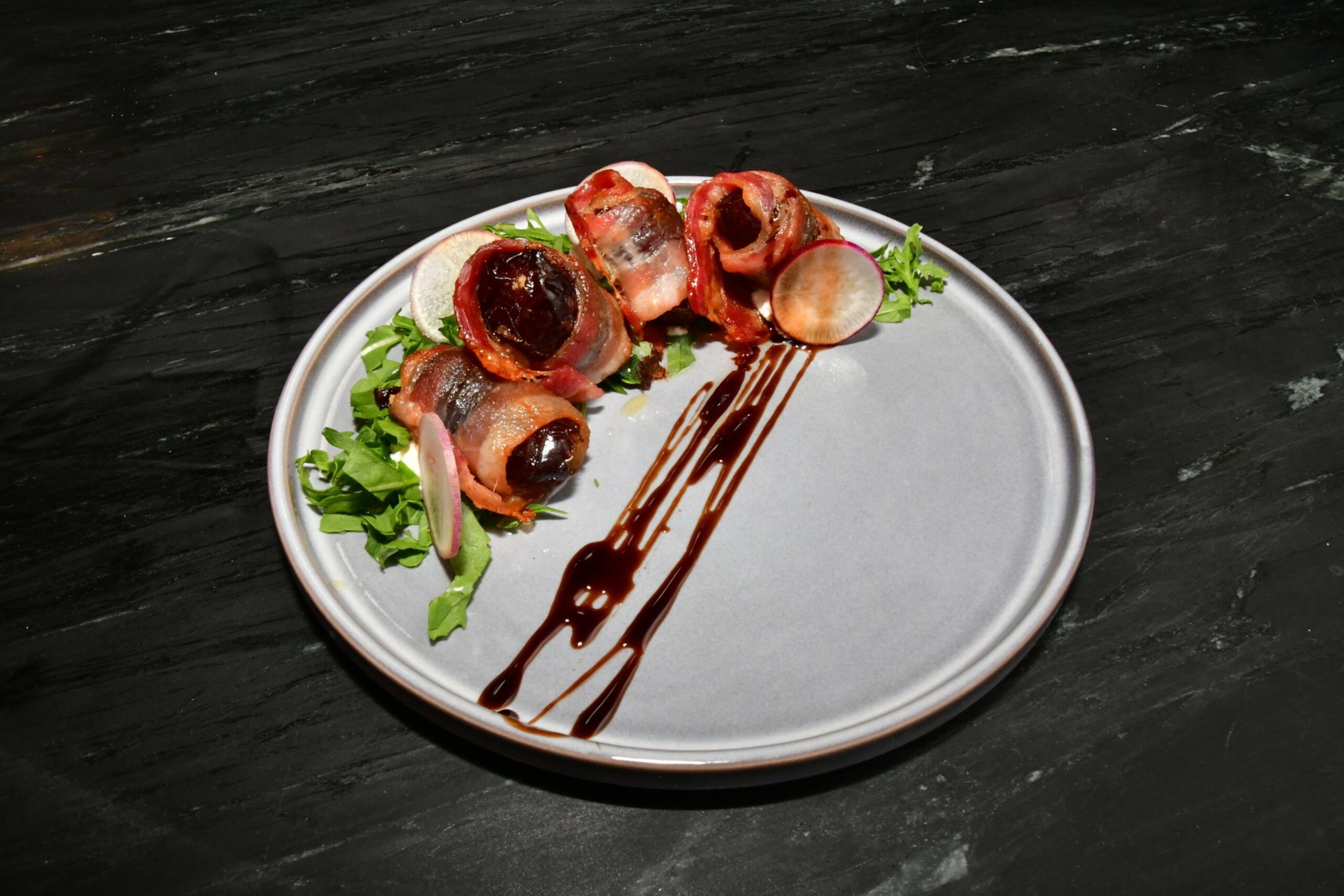 Bacon-wrapped dates are arranged on greens with radish slices, alongside streaks of balsamic glaze on a gray plate over a black surface.
