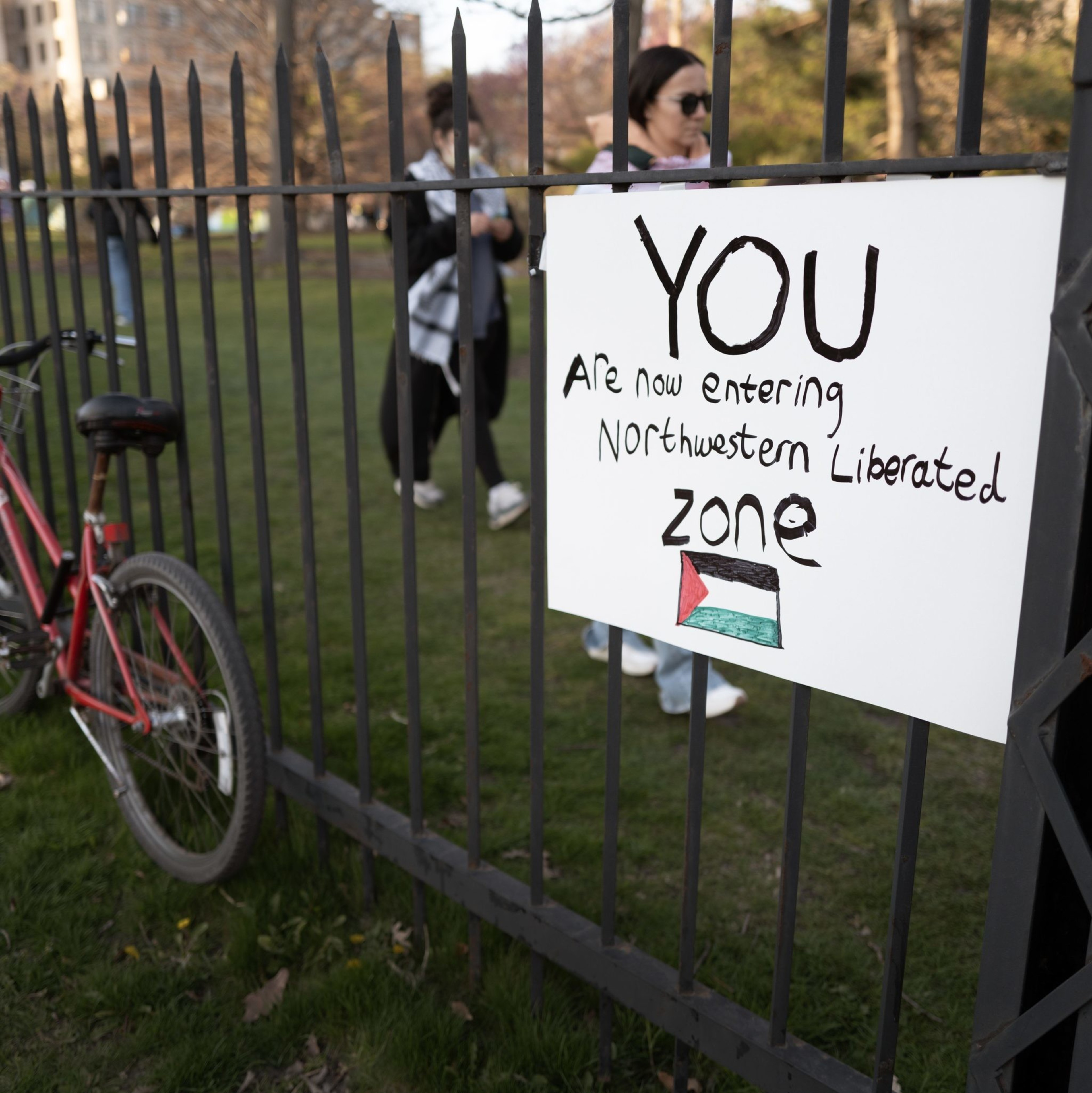 A sign on a metal fence reads, &quot;You are now entering Northwestern Liberated zone,&quot; with a Palestinian flag drawn below it. A red bike is also secured to the fence.