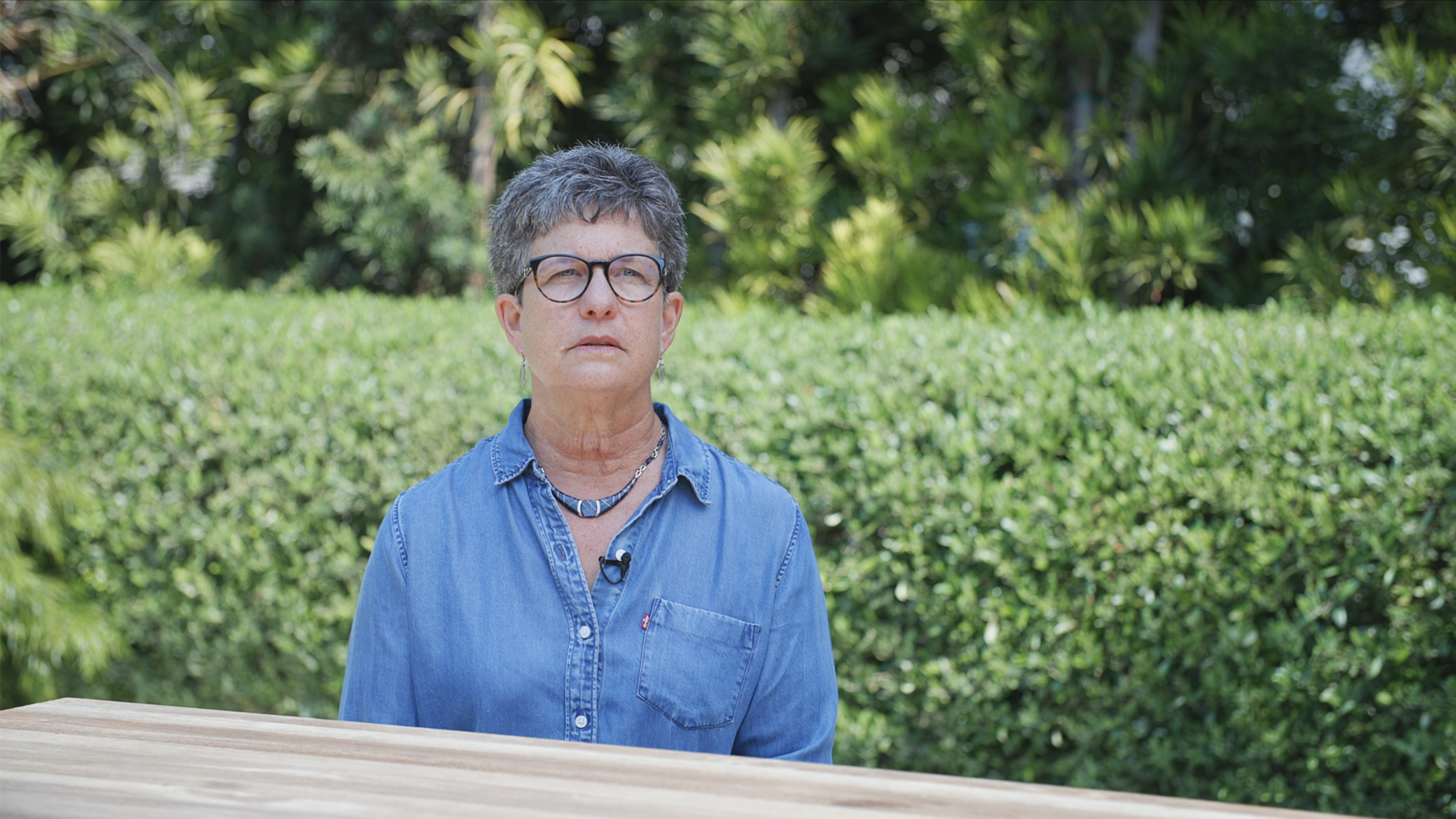 A person with short gray hair and glasses wears a blue denim shirt and a necklace, sitting outdoors with green bushes in the background.