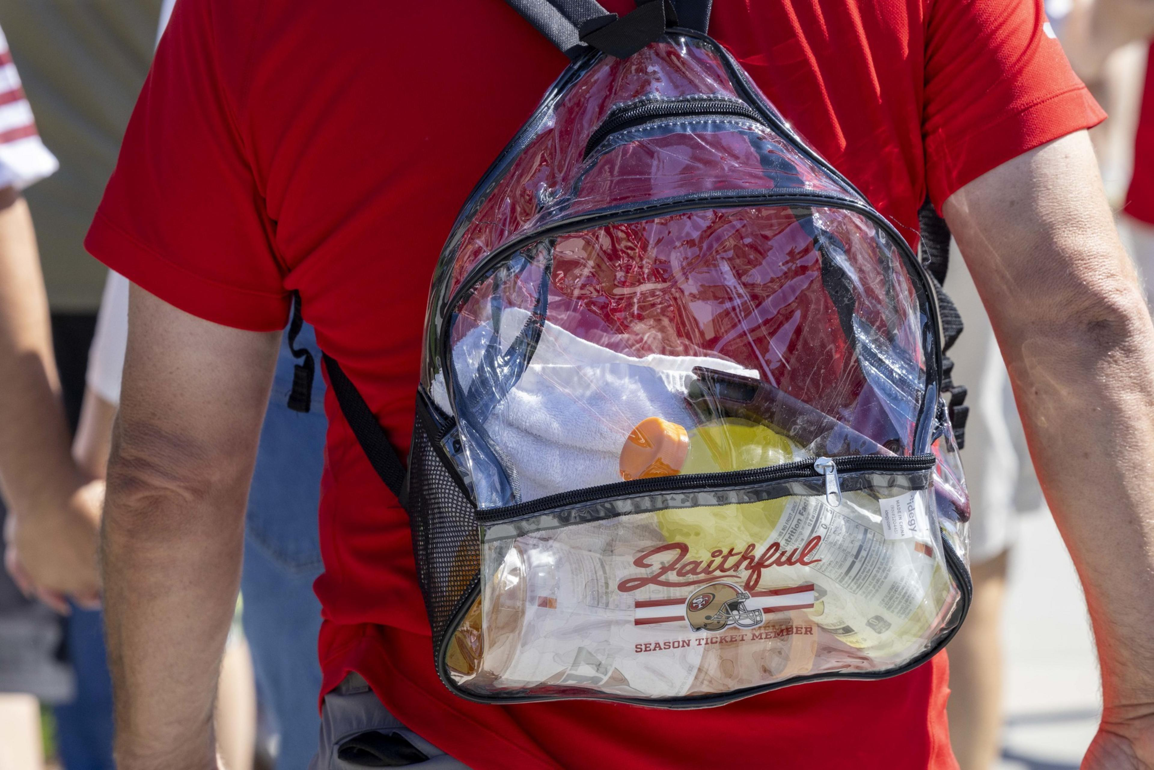 A person in a red shirt is wearing a clear backpack, revealing a water bottle, towel, and items labeled &quot;Faithful&quot; with a sports team logo.