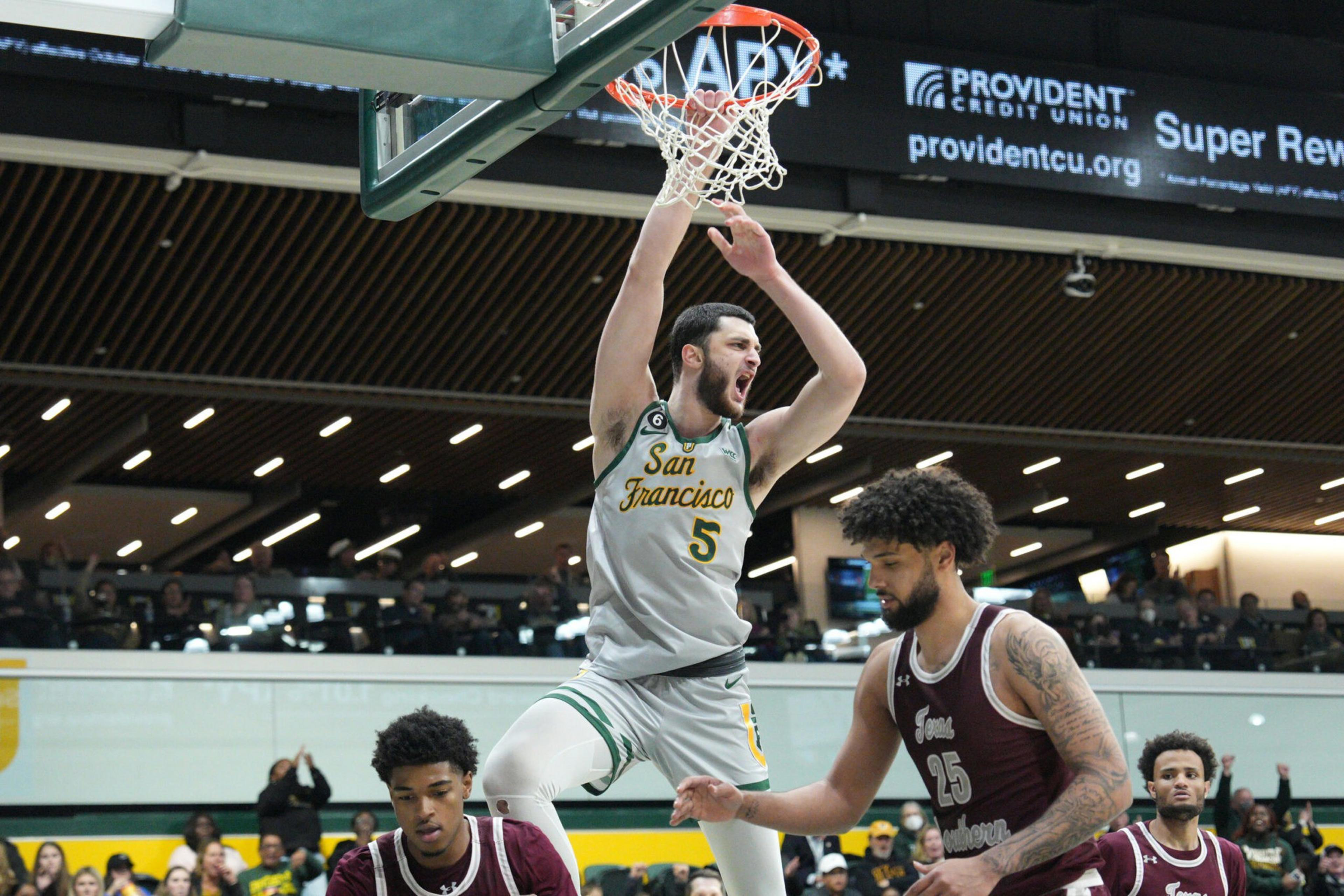 A basketball player in a gray "San Francisco" jersey dunks the ball while two opponents in maroon jerseys watch below.