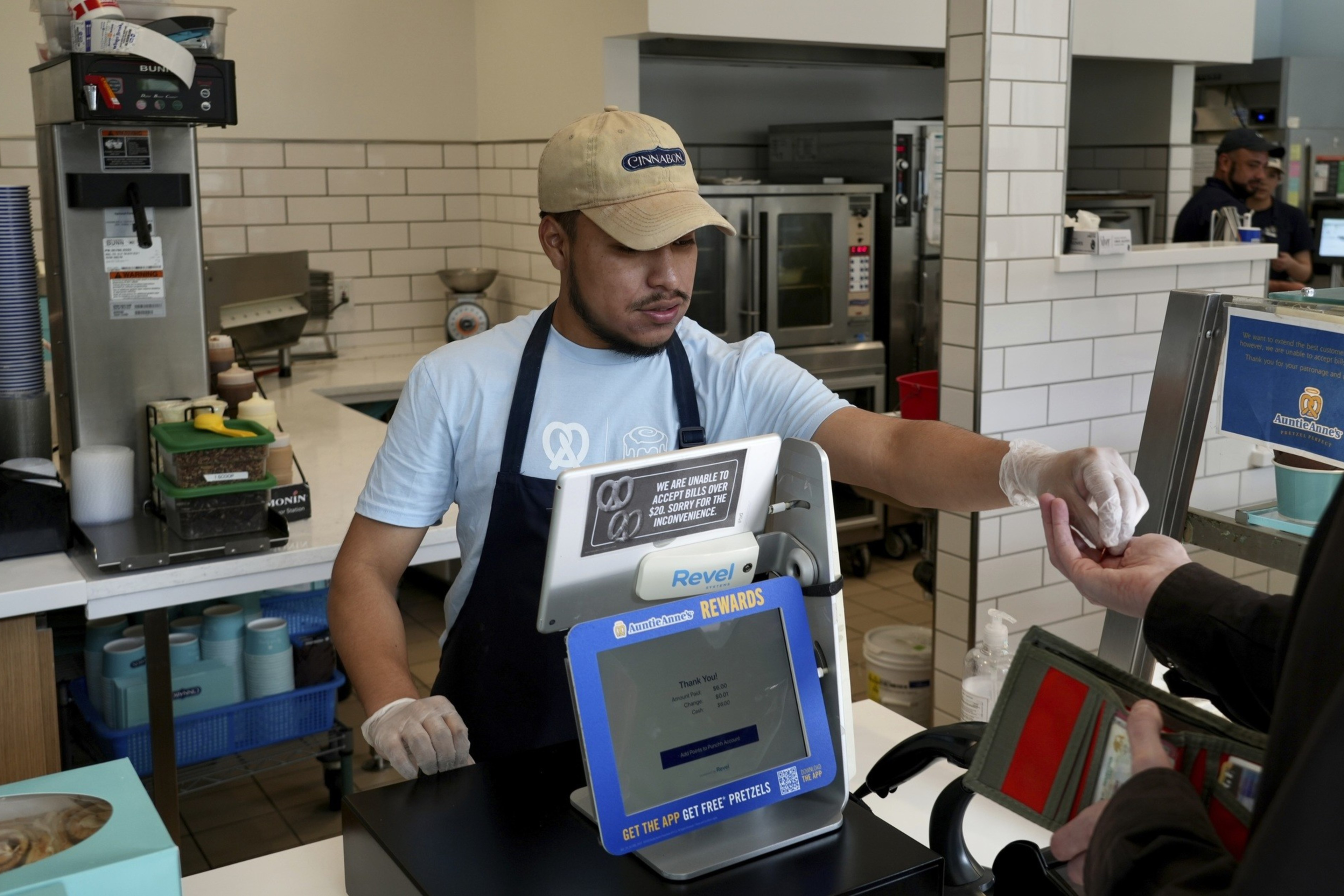 A cashier in a cap and apron hands a receipt to a customer at a fast-food counter, with kitchen equipment in the background.