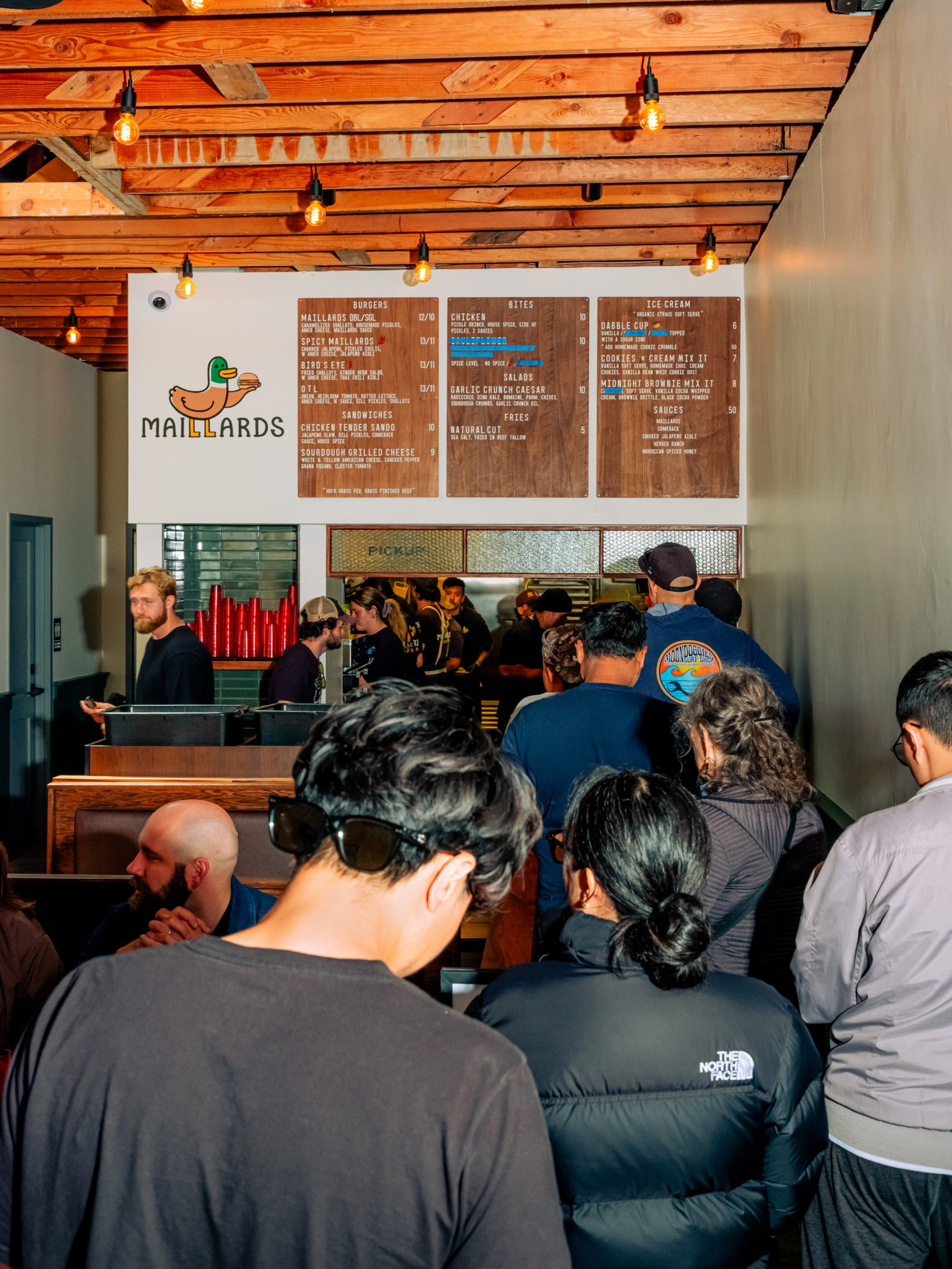 A crowd of people stands inside a casual eatery with a wooden ceiling, facing a wall menu featuring burgers, sandwiches, salads, and ice cream at Maillards.
