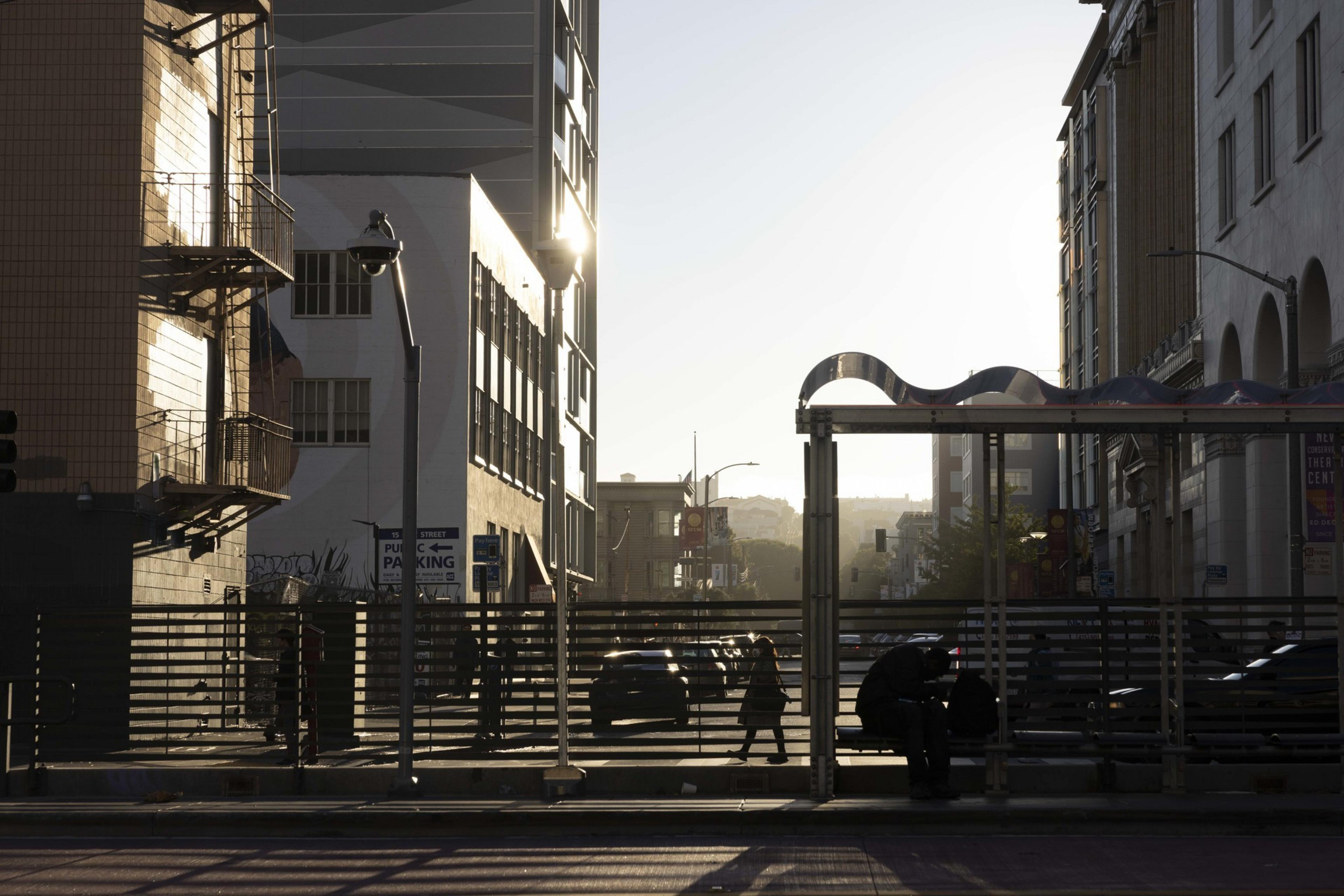 The image shows a city street at dawn or dusk with a golden light casting shadows. There are buildings, a streetlight, people walking, and a bus stop in silhouette.