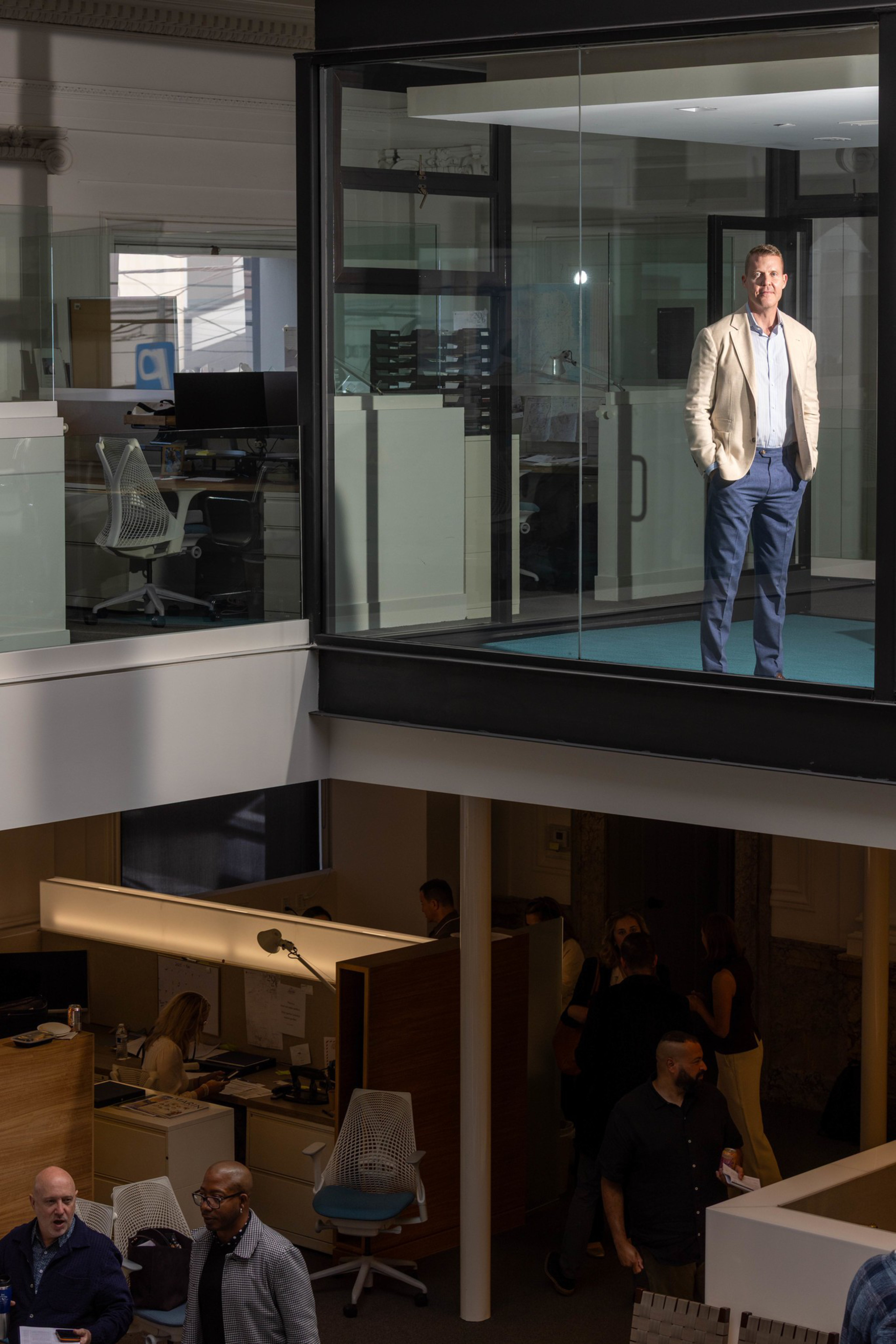 A man in a beige jacket and blue pants stands alone in a glass-walled office above, while several people work and talk in a darker area below.