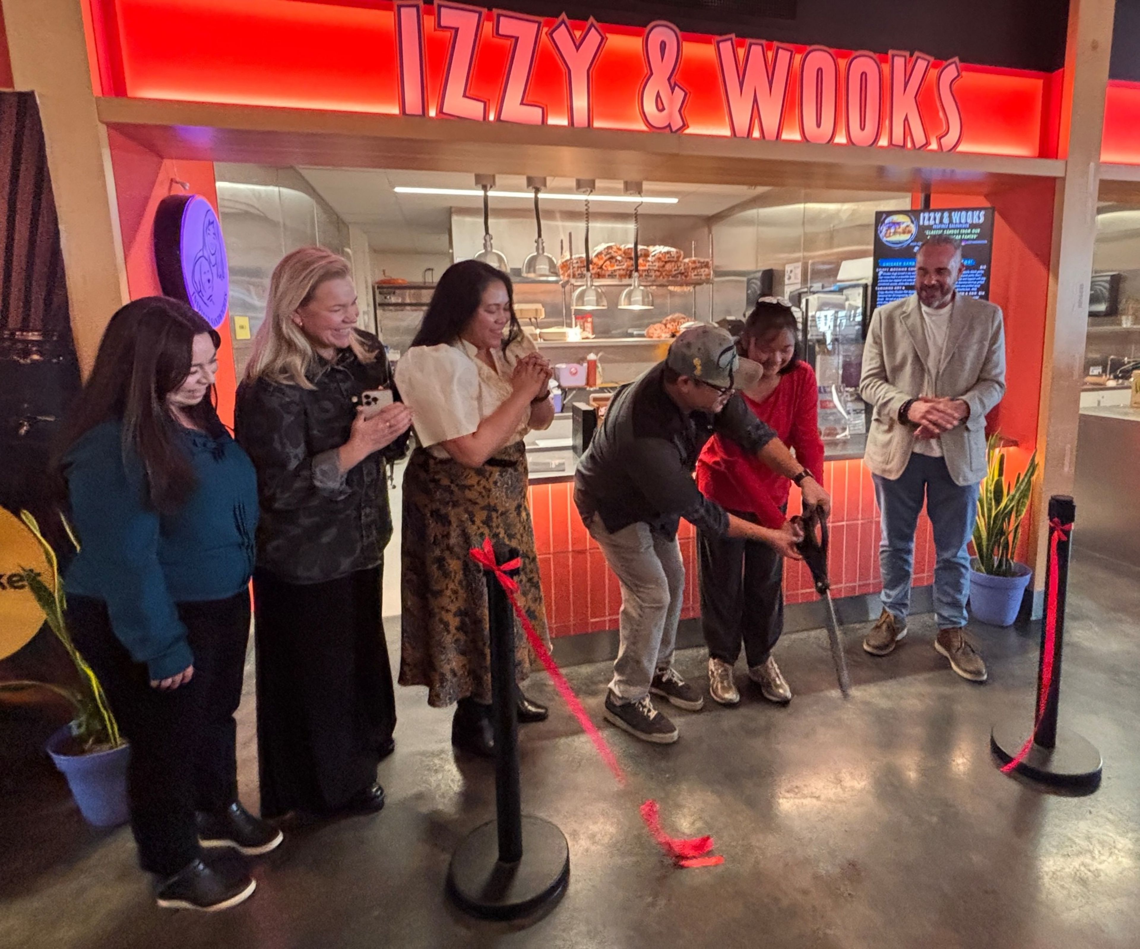 Six people smile as one cuts a red ribbon in front of a brightly lit Izzy & Wooks restaurant entrance, marking a celebratory opening event.