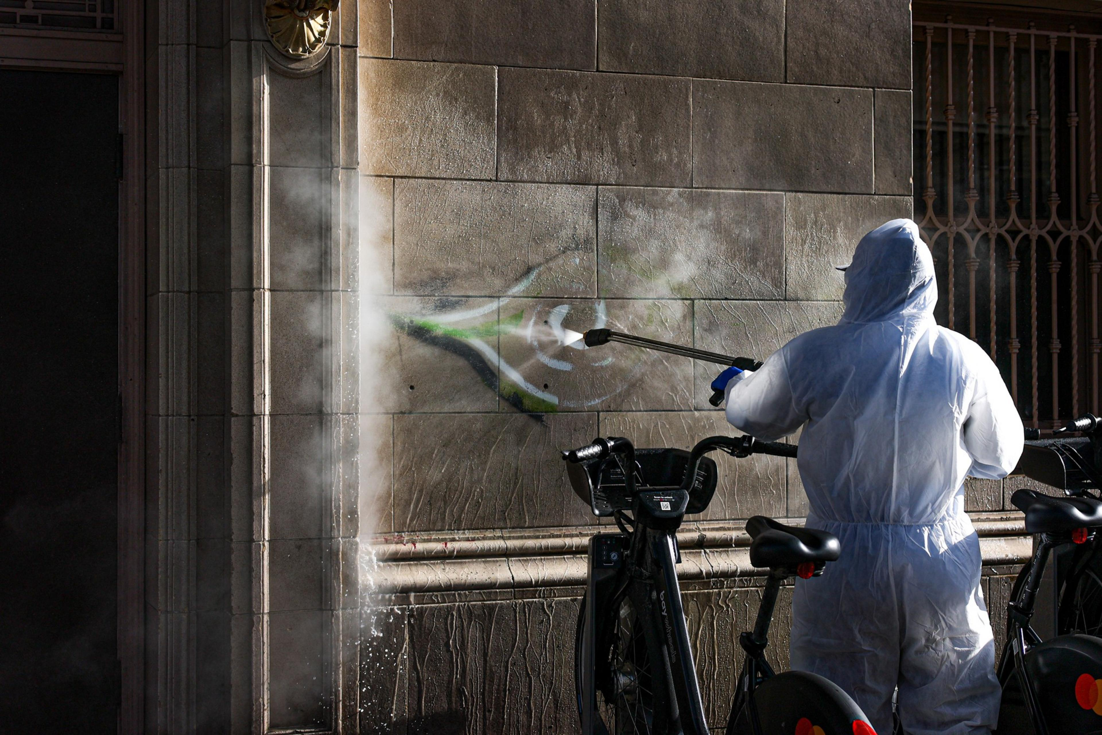 A person in a white protective suit pressure washes graffiti off a stone wall near parked bikes in sunlight.