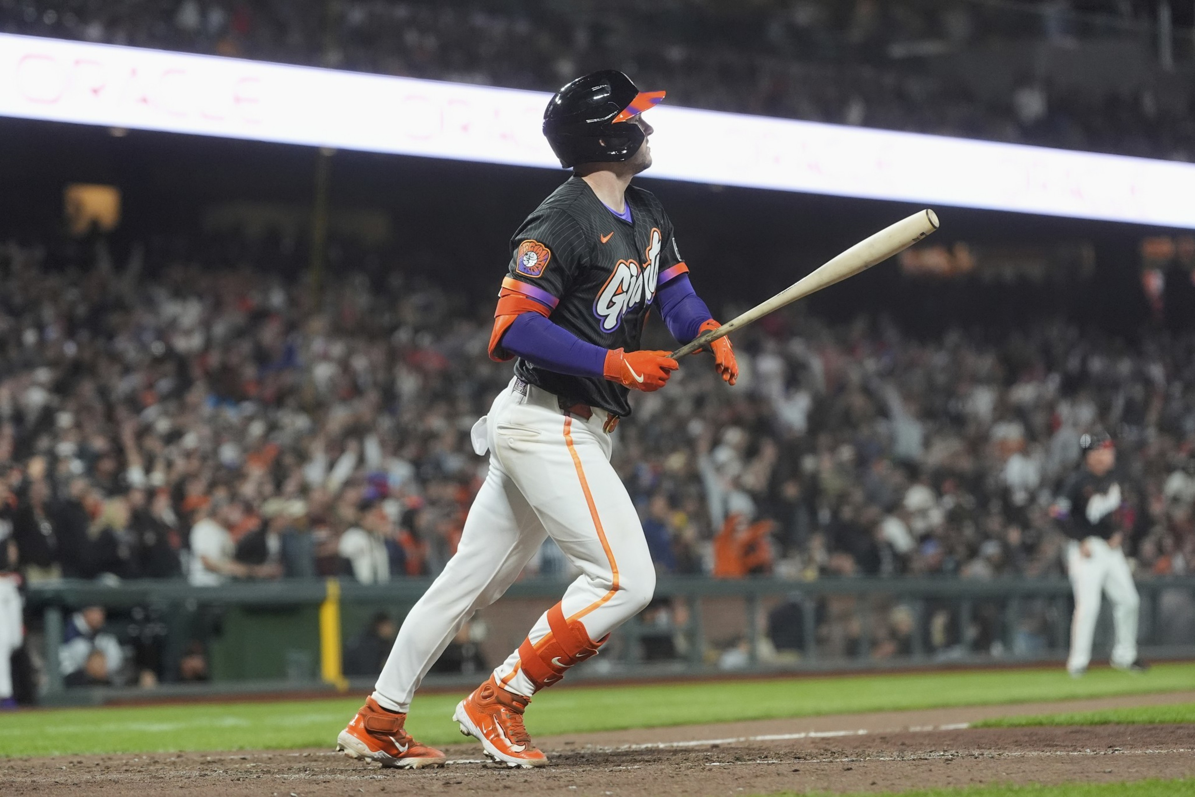 A baseball player in a black and orange uniform watches the ball after hitting it, standing on a field with a cheering crowd in the background.