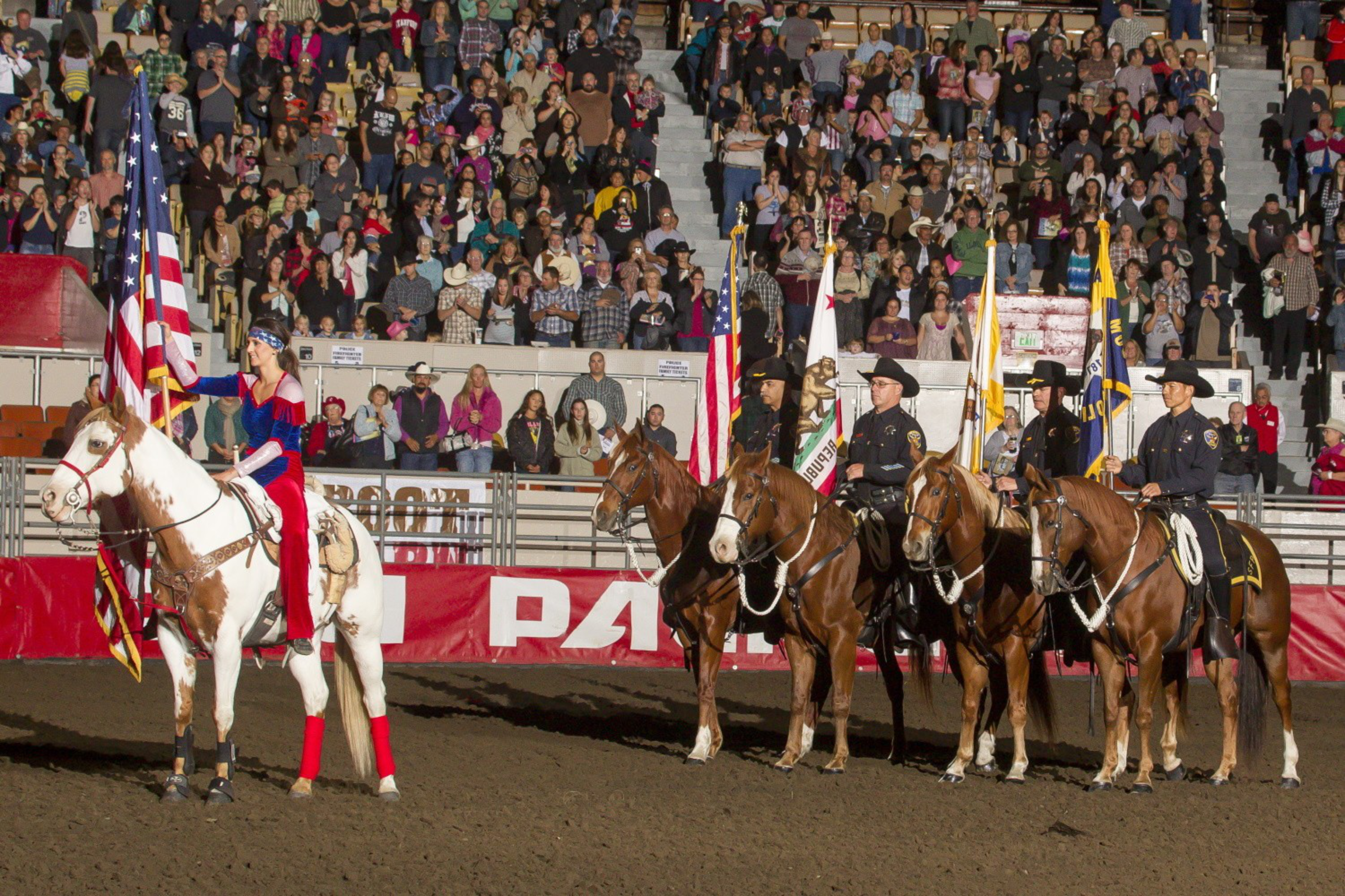 Riders on horseback carry flags in an arena, with spectators watching in the background.