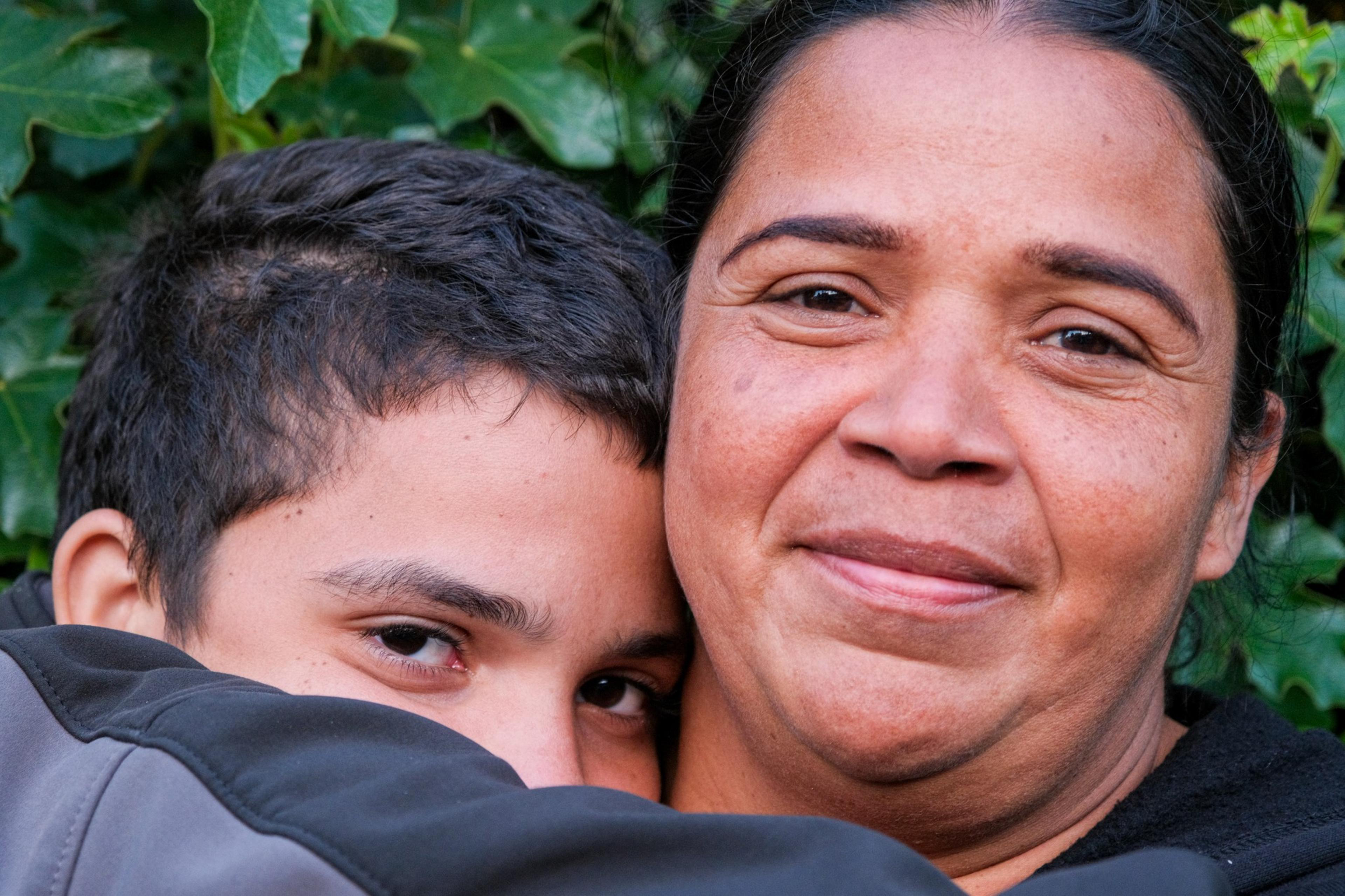 A boy is hugging a smiling woman; both have dark hair, and ivy leaves form the background.
