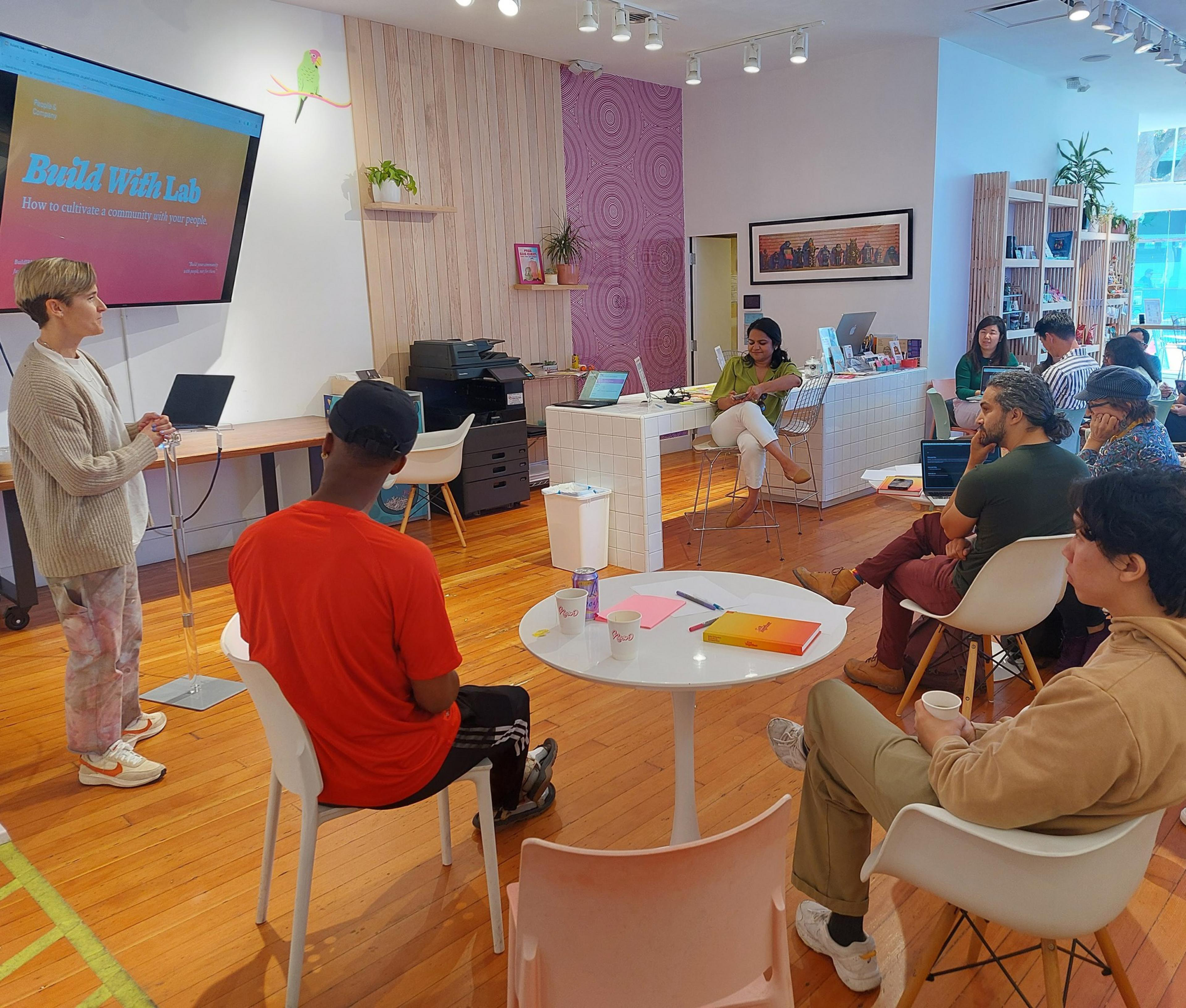 A person presents to a small group in a modern, brightly lit room with wooden floors. Attendees sit around tables with laptops, notepads, and drinks.