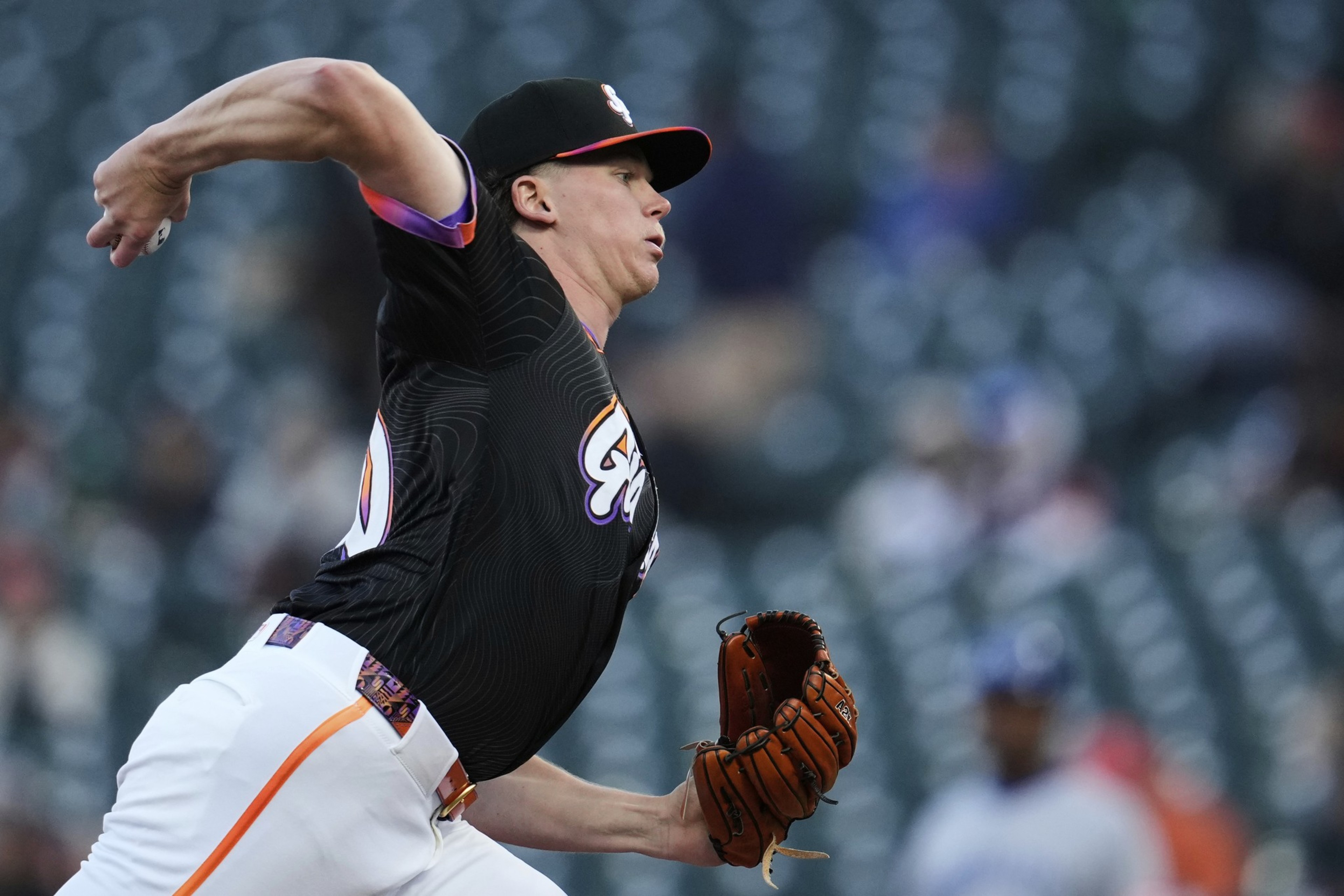 A baseball player in a black jersey is pitching, with his right arm extended and a glove in his left hand. He is in a stadium with empty green seats behind him.
