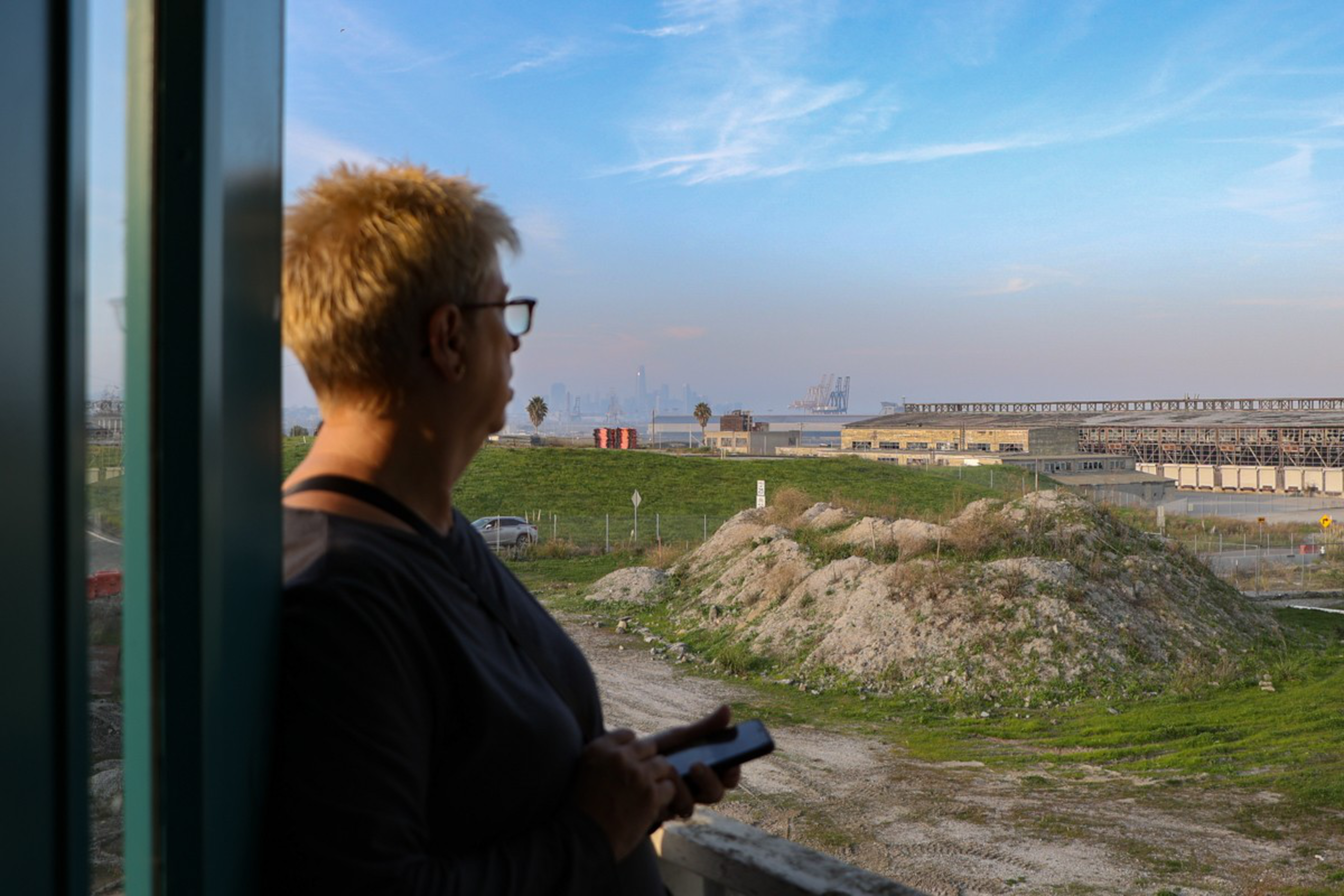 A person with short blond hair and glasses looks out over a dirt mound and green grass toward industrial buildings and a hazy city skyline under a blue sky.